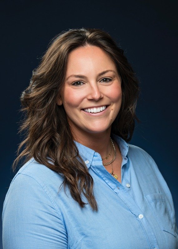 Portrait of a woman with long wavy brown hair, smiling, wearing a light blue button-up shirt, against a dark background.