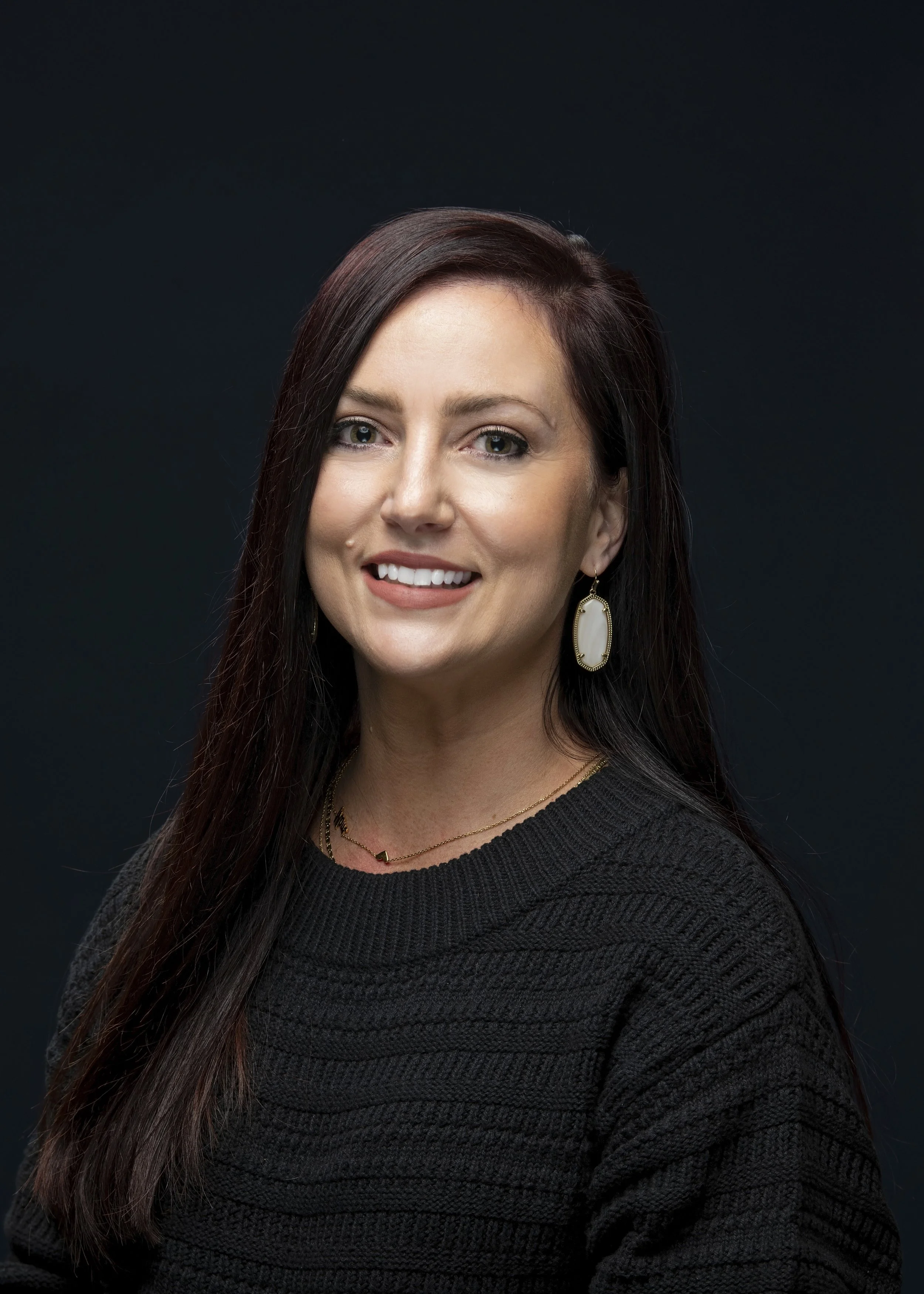 Portrait of a woman with long dark hair, smiling at the camera, wearing a black top, earrings, and a necklace, against a dark background.