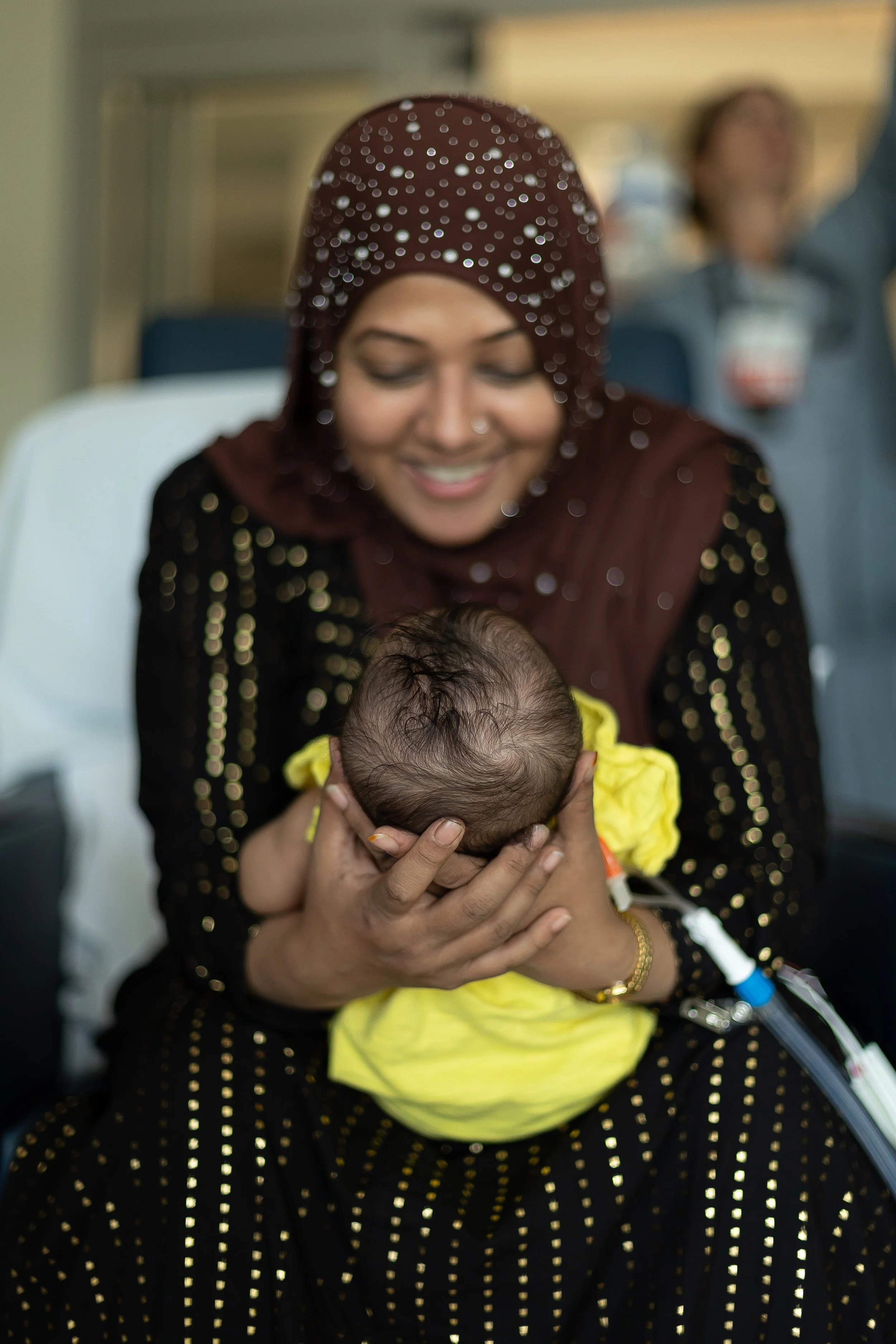 A woman wearing a maroon and black dress with gold dots and a maroon hijab with sequins, sitting in a hospital bed, holding a newborn baby with a bandage on its head and a yellow blanket, smiling and looking down at the baby.