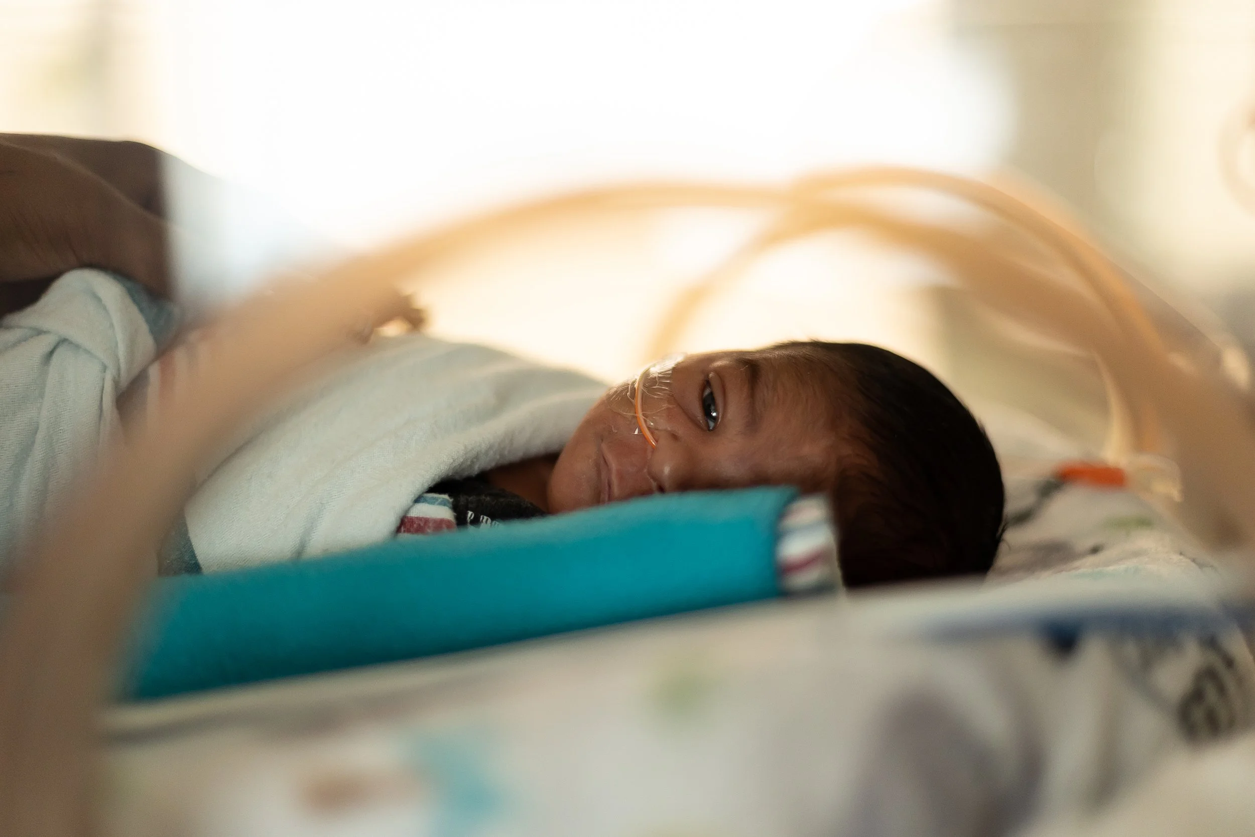A young child lying in a hospital bed, connected to a breathing tube, with a blanket covering them.