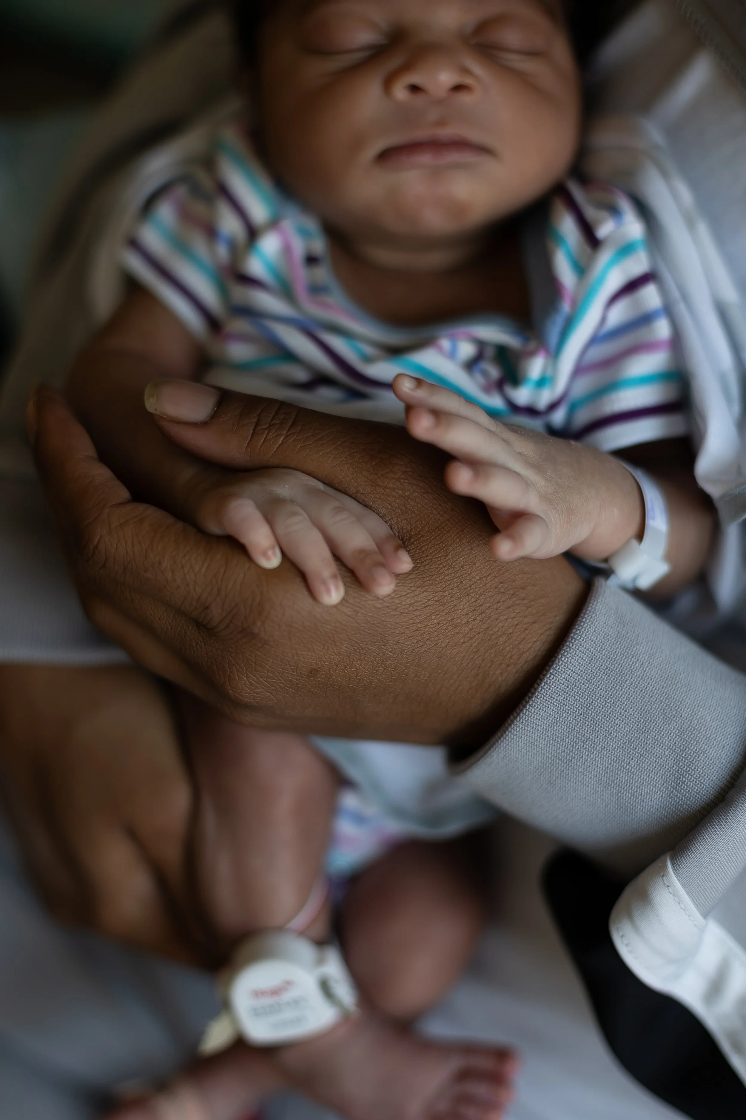 Close-up of a sleeping baby gently holding a person's hand, in a hospital setting.