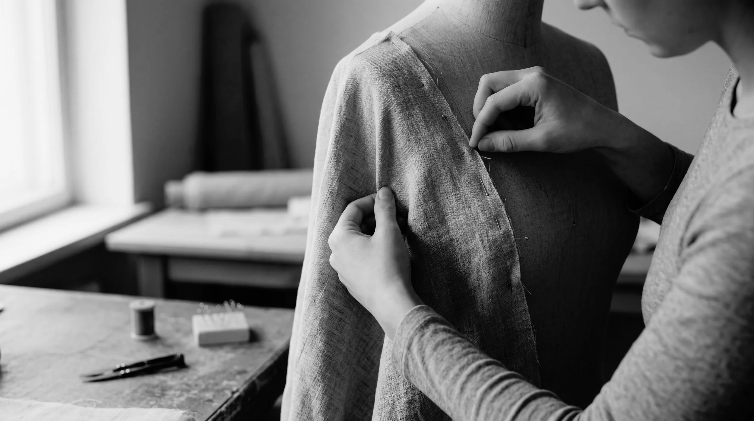 Hands adjusting linen fabric on a dress form mannequin in a tailoring studio