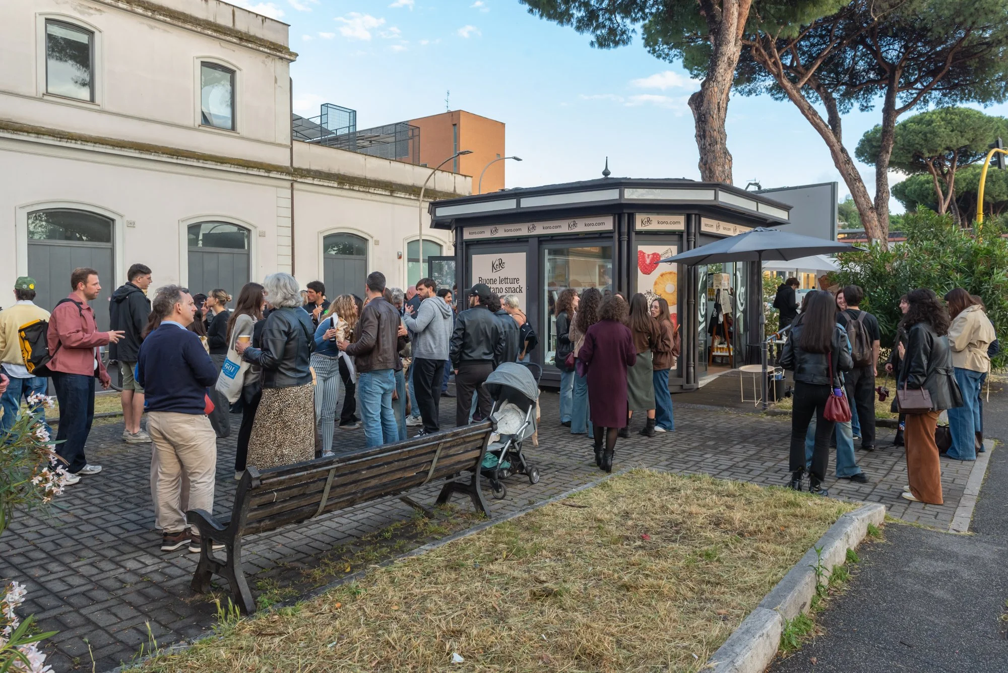 Gruppo di persone che si riuniscono e socializzano all'aperto, attorno a una cabina in cui si vendono snack e libri. Alcuni sono in piedi, altri seduti su una panchina. L'atmosfera sembra informale e vivace, in un'area verde con alberi e piante.
