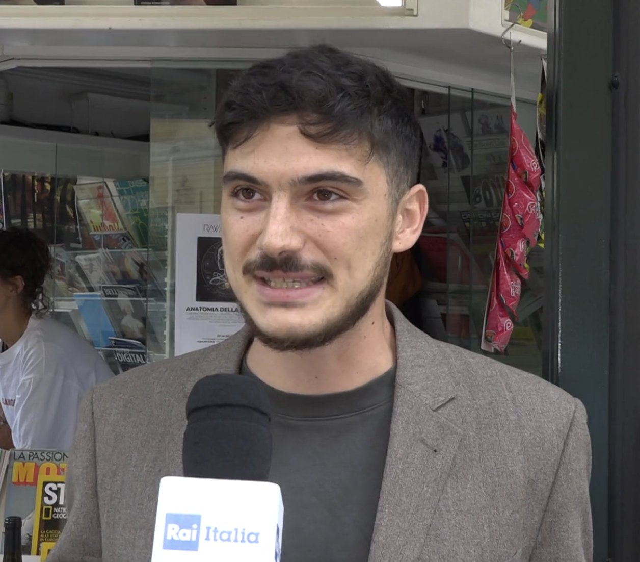 Un giovane uomo con barba, capelli castani e vestito con una giacca marrone, sta parlando davanti a un microfono con il logo Rai Italia, in un contesto urbano o commerciale, con una libreria e alcune decorazioni sullo sfondo.