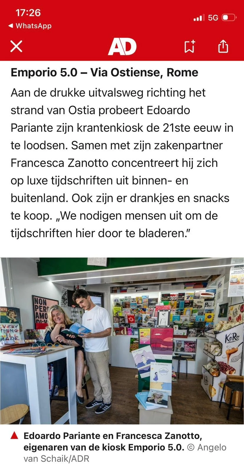 immagine di una libreria con due persone, una donna seduta e un uomo che le mostra un libro, scaffali pieni di libri e riviste sullo sfondo
