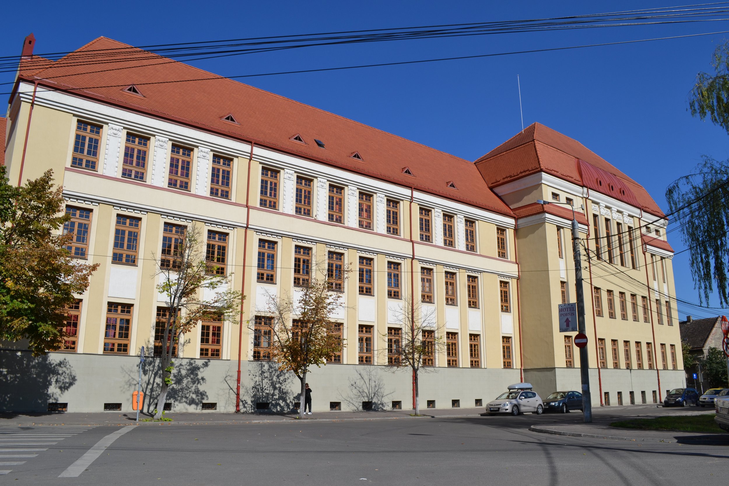 Large beige building with red roof and multiple windows, situated by the street with cars parked along the curb and trees casting shadows.