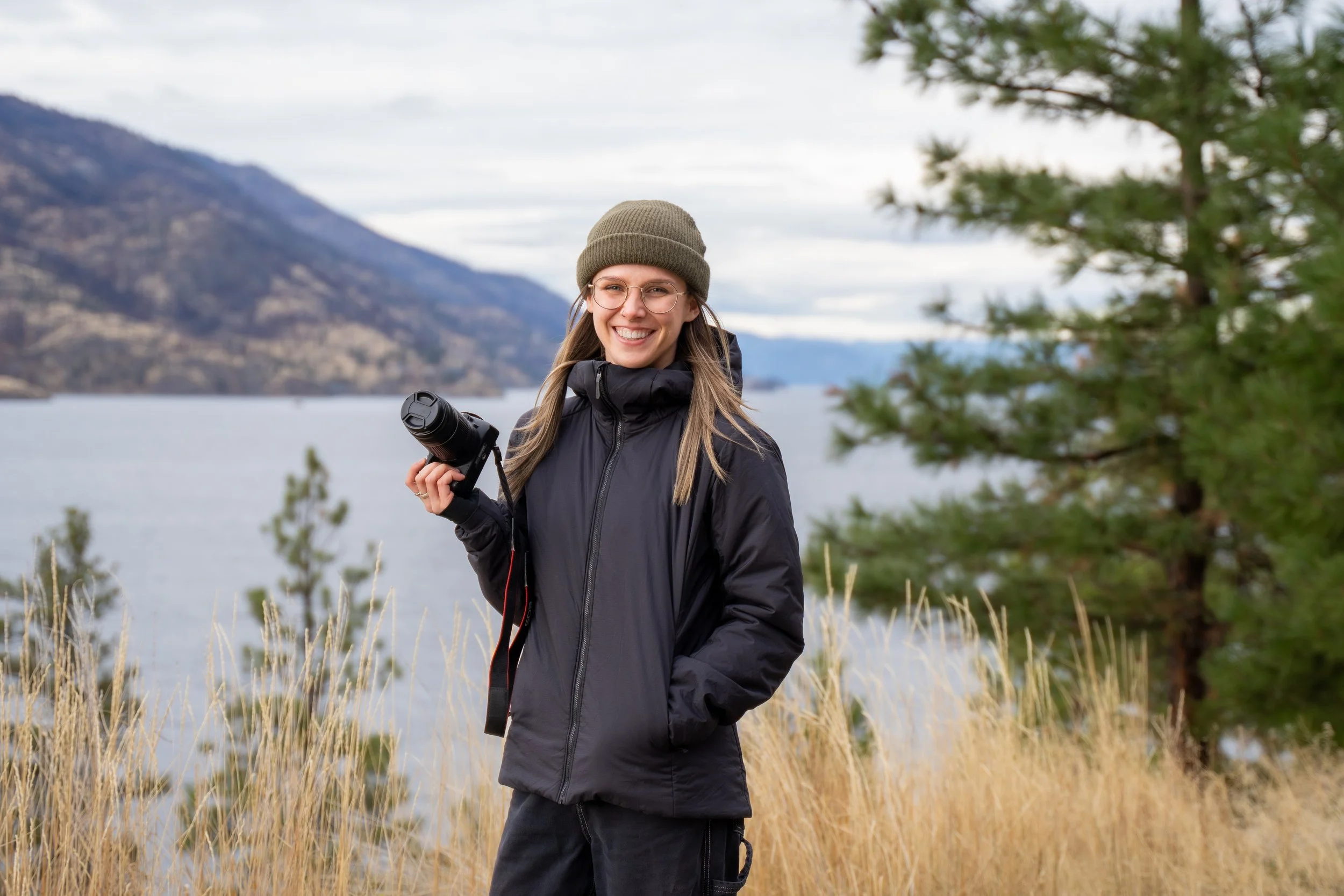 A smiling young woman in outdoor gear holding a camera stands near a body of water with mountains and pine trees in the background.
