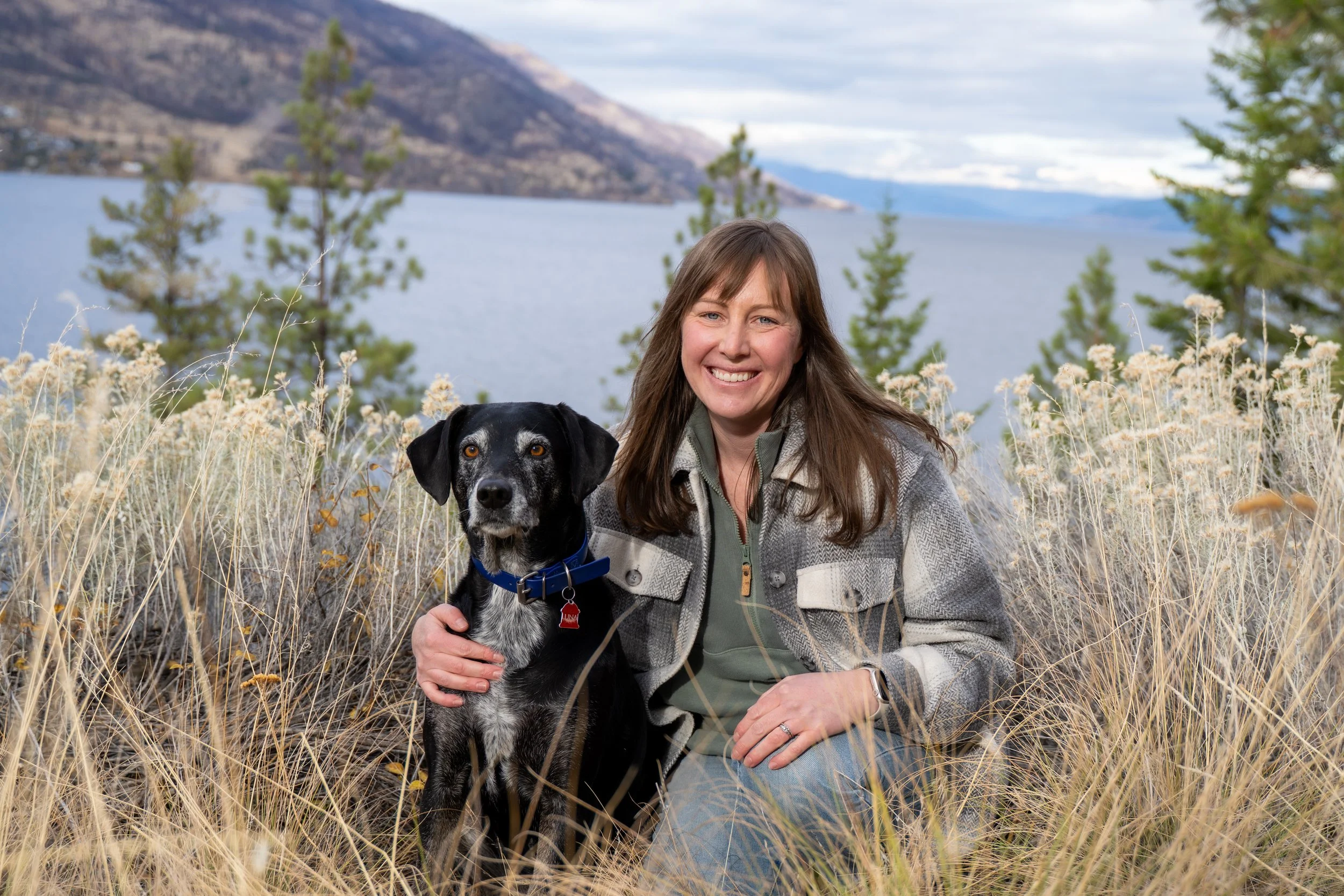 A woman smiling with a dog in a grassy field near a lake with mountains in the background.