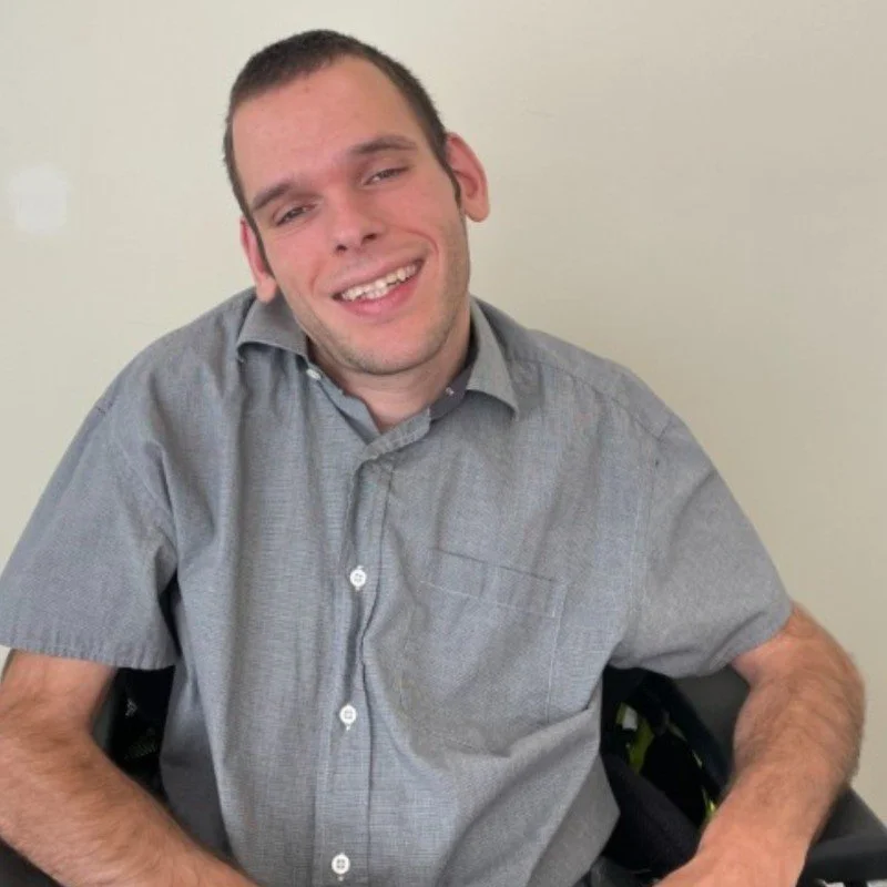 A man with curly dark hair, glasses, a beard and mustache, and a small nose ring, wearing a dark green sweater and a white shirt underneath, sitting indoors with a neutral background.
