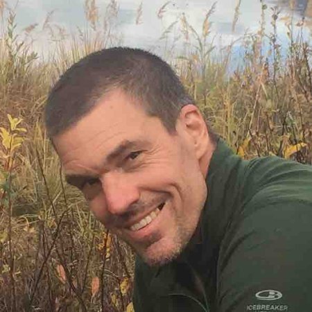 A woman with curly black hair, wearing glasses, hoop earrings, multiple necklaces, and a white striped shirt, smiling.