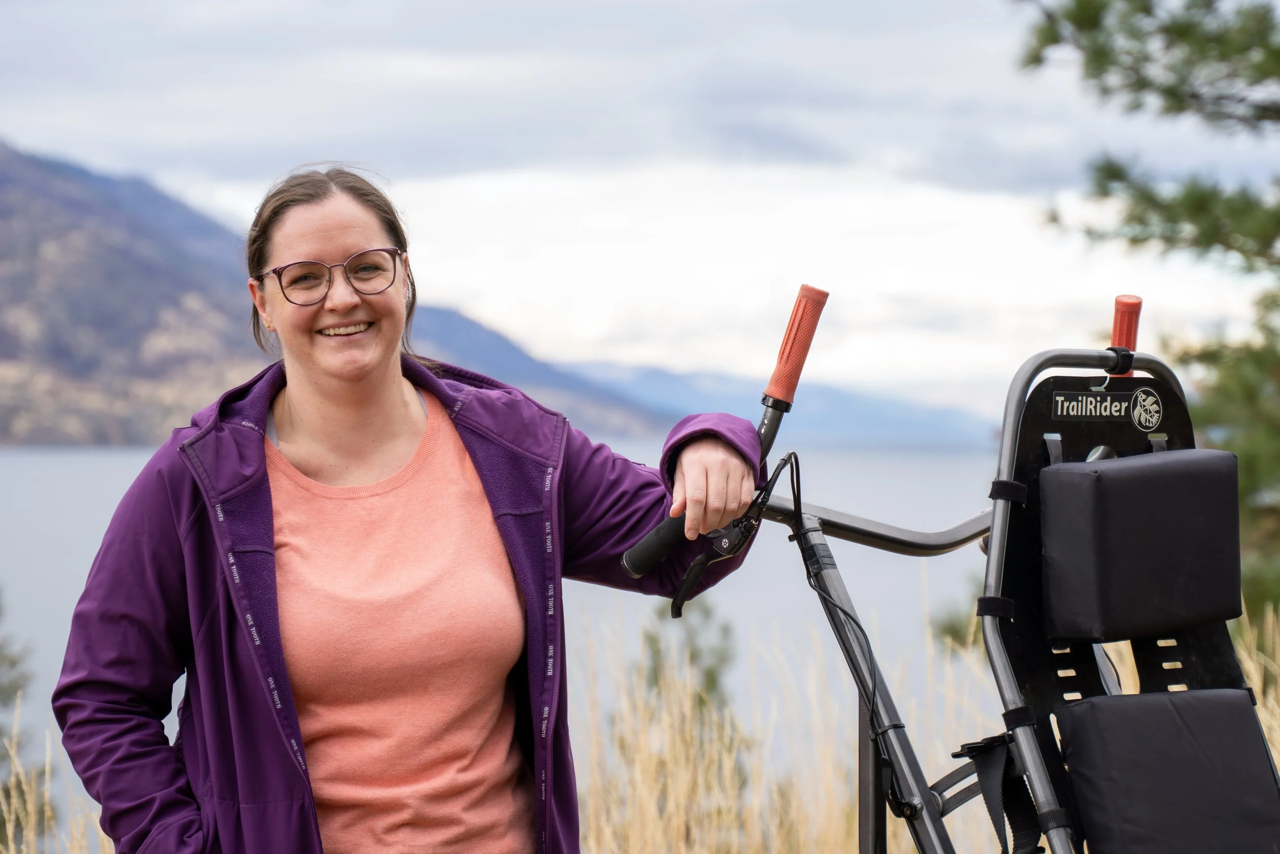 A woman with glasses and a purple jacket smiling while standing outdoors near a lake, holding a trekking stroller.