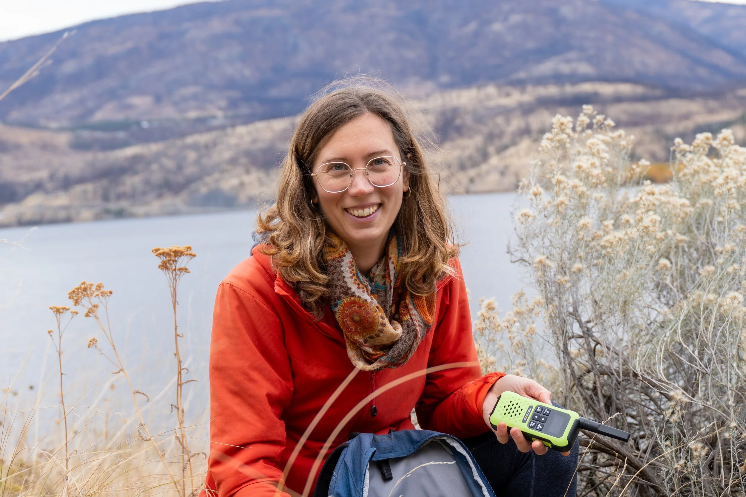 A woman with glasses and wavy brown hair smiling outdoors in front of a lake and mountains, wearing an orange jacket and holding a portable radio.