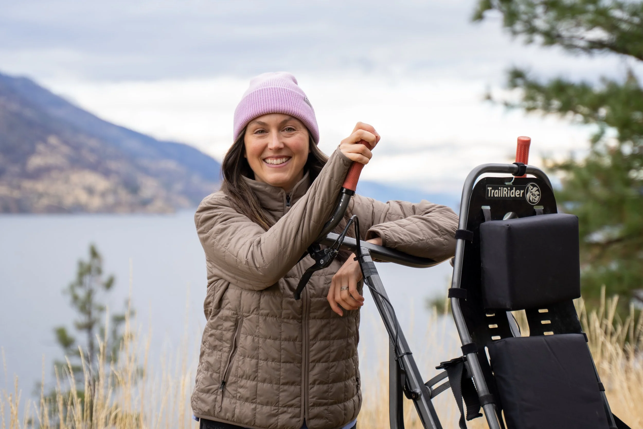 A man with curly dark hair, glasses, and a beard. He is wearing a black sweater and looking slightly to the side with a thoughtful expression. There is a blurred plant and a light-colored wall in the background.