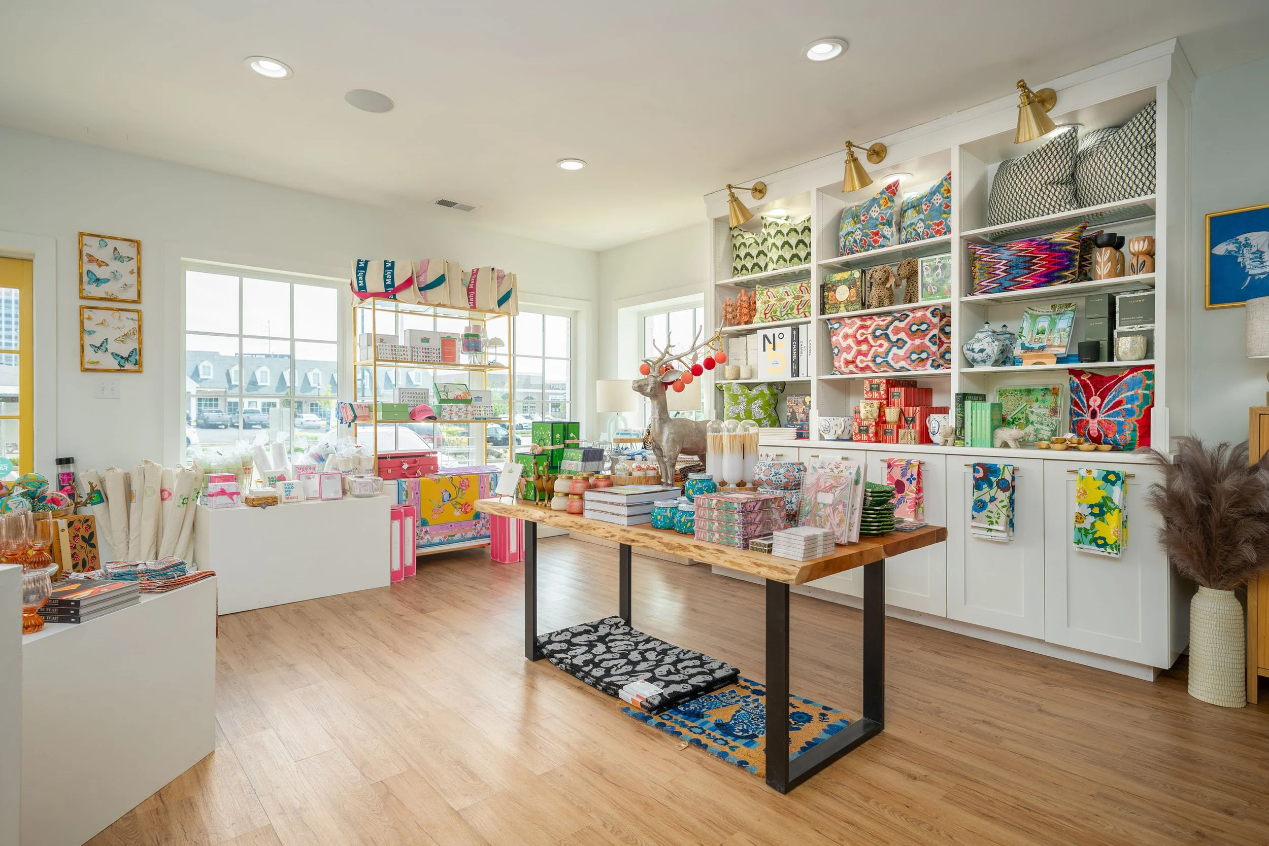 Bright retail store with shelves of colorful home decor and accessories, a central wooden table with decorative boxes, a reindeer sculpture, and large windows letting in natural light.