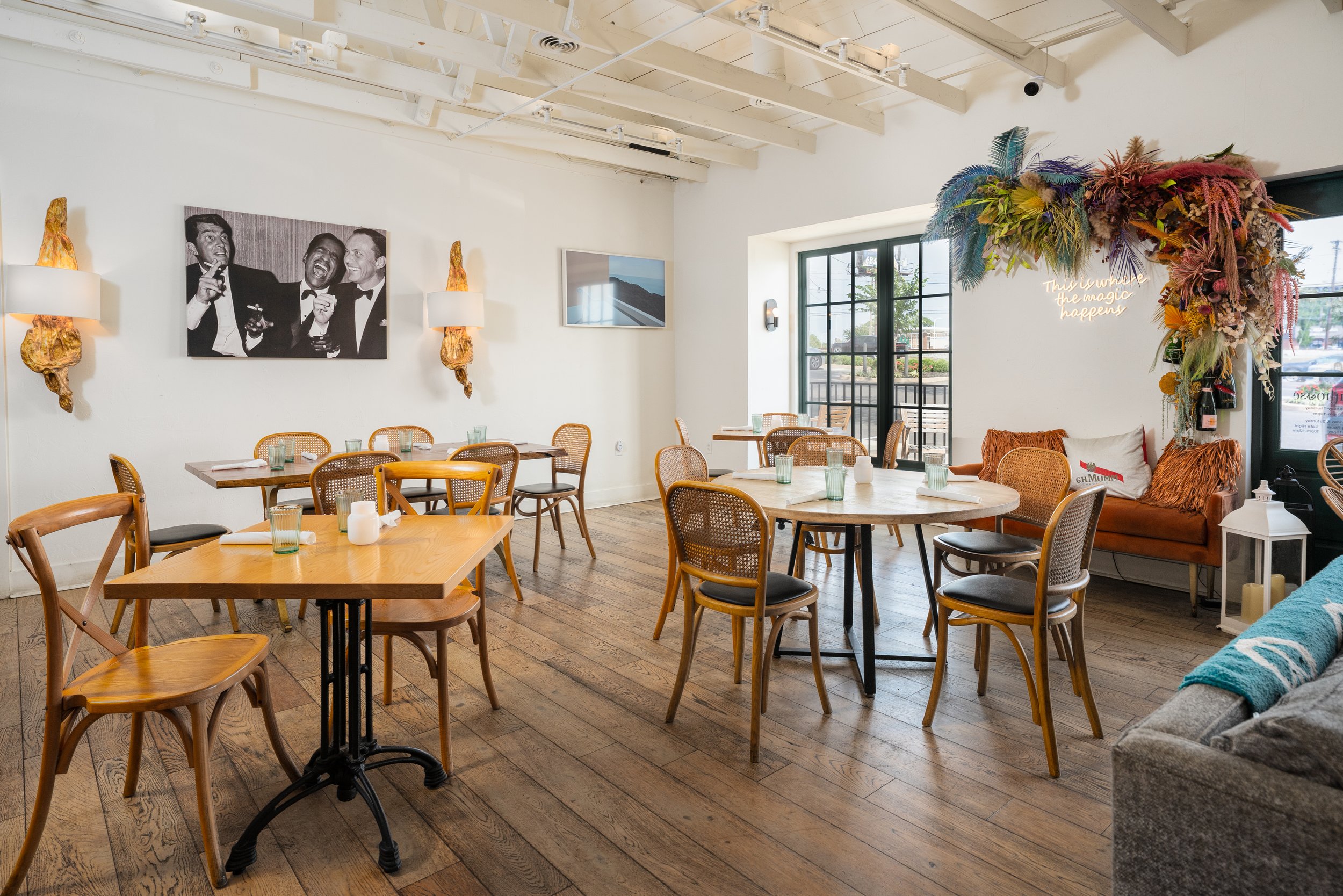 Interior of a bright, coastal-style restaurant with wooden flooring, white walls, and large windows. There are various tables and chairs, a patterned orange sofa, wall art including a black-and-white photo, and a colorful floral and feather wall deco