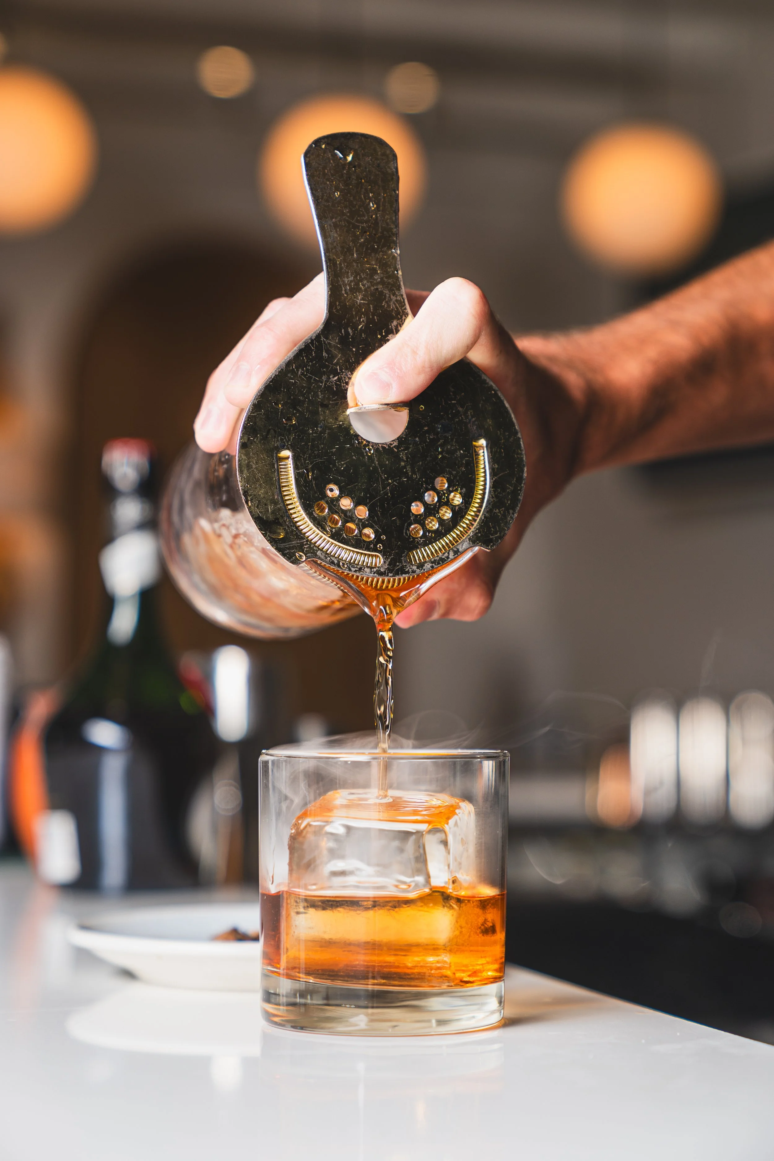 A bartender pours whiskey from a cocktail shaker into a glass with a large ice cube.