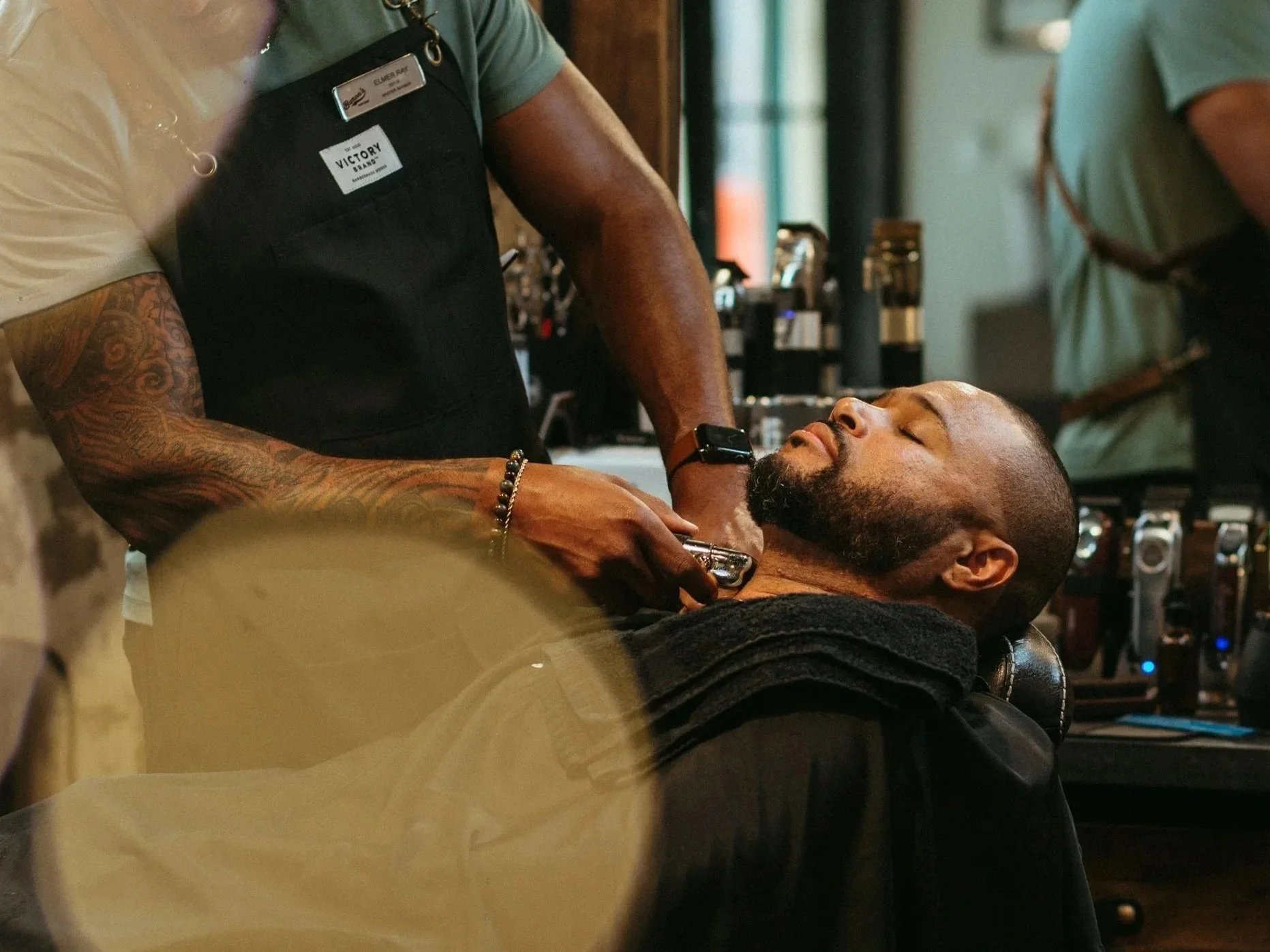 A man is getting a shave at a barbershop by a barber with tattoos on his arm, and the man appears relaxed with eyes closed.