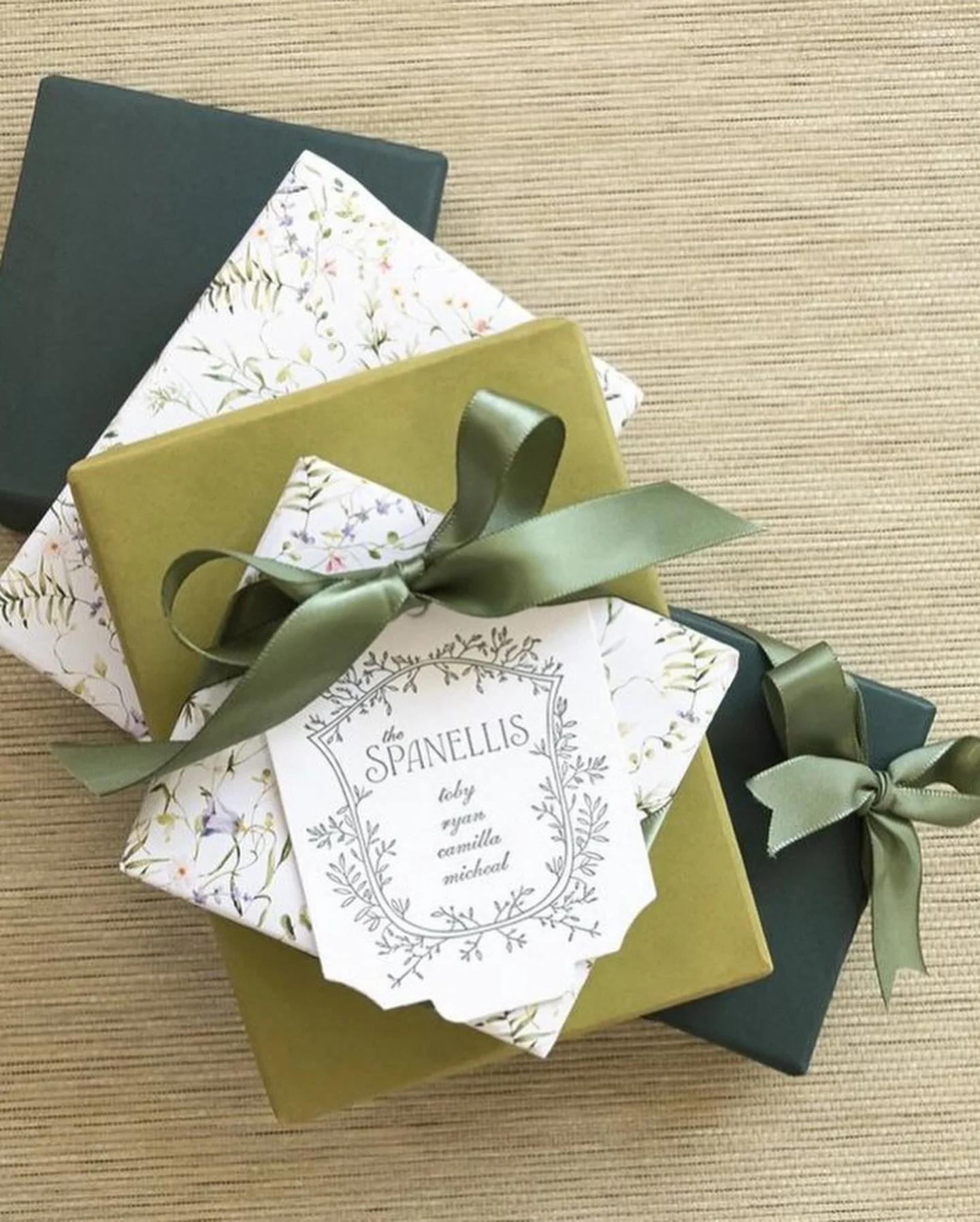 Three wrapped gift boxes with floral and solid wrapping paper, adorned with olive green ribbons, placed on a striped beige surface.