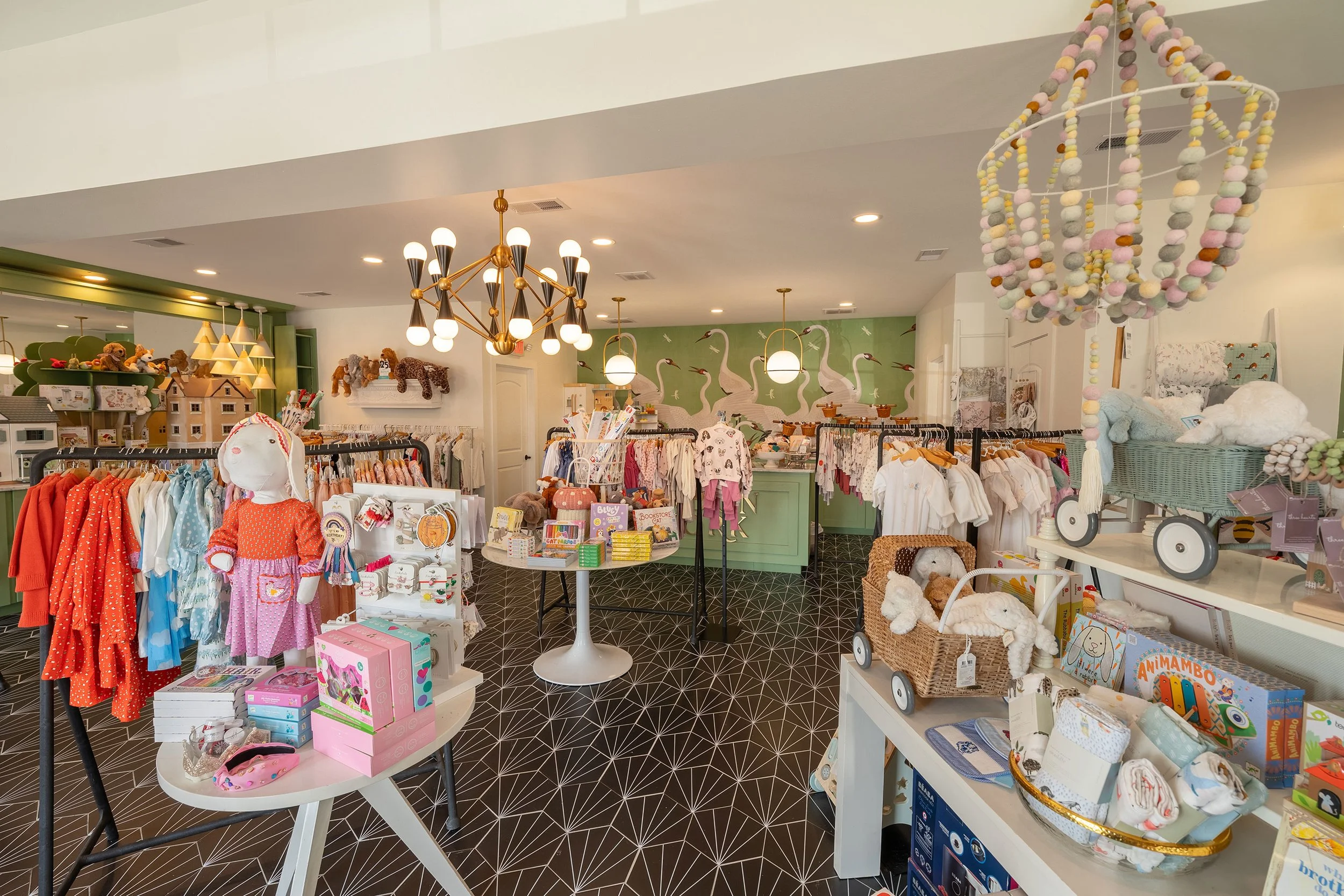 Interior of a children's boutique store decorated with plush toys, children’s clothing, and accessories, featuring a green mural of swans on the back wall and unique lighting fixtures.