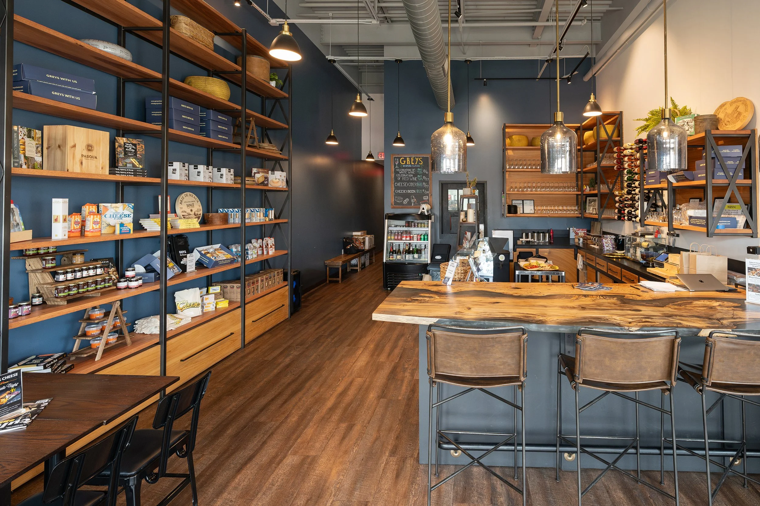 Interior of a modern store or café with wooden shelves stocked with products, a wooden counter with chairs, and a dark blue wall with a chalkboard menu. The space features industrial-style lighting and a wood floor.