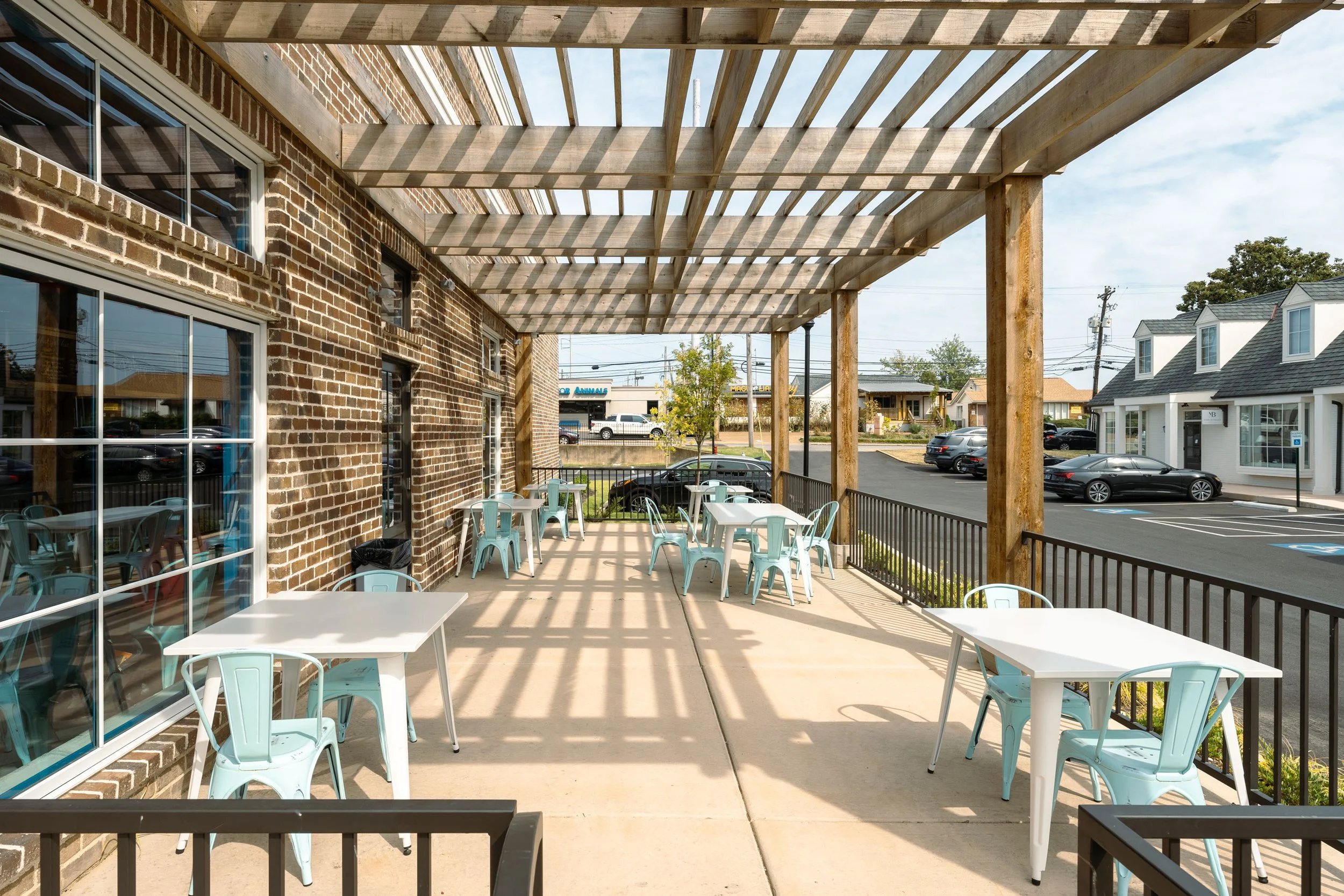 Outdoor patio with white tables and light blue chairs, covered by a wooden pergola, adjacent to a brick building, overlooking a parking lot and neighboring houses under a clear sky.
