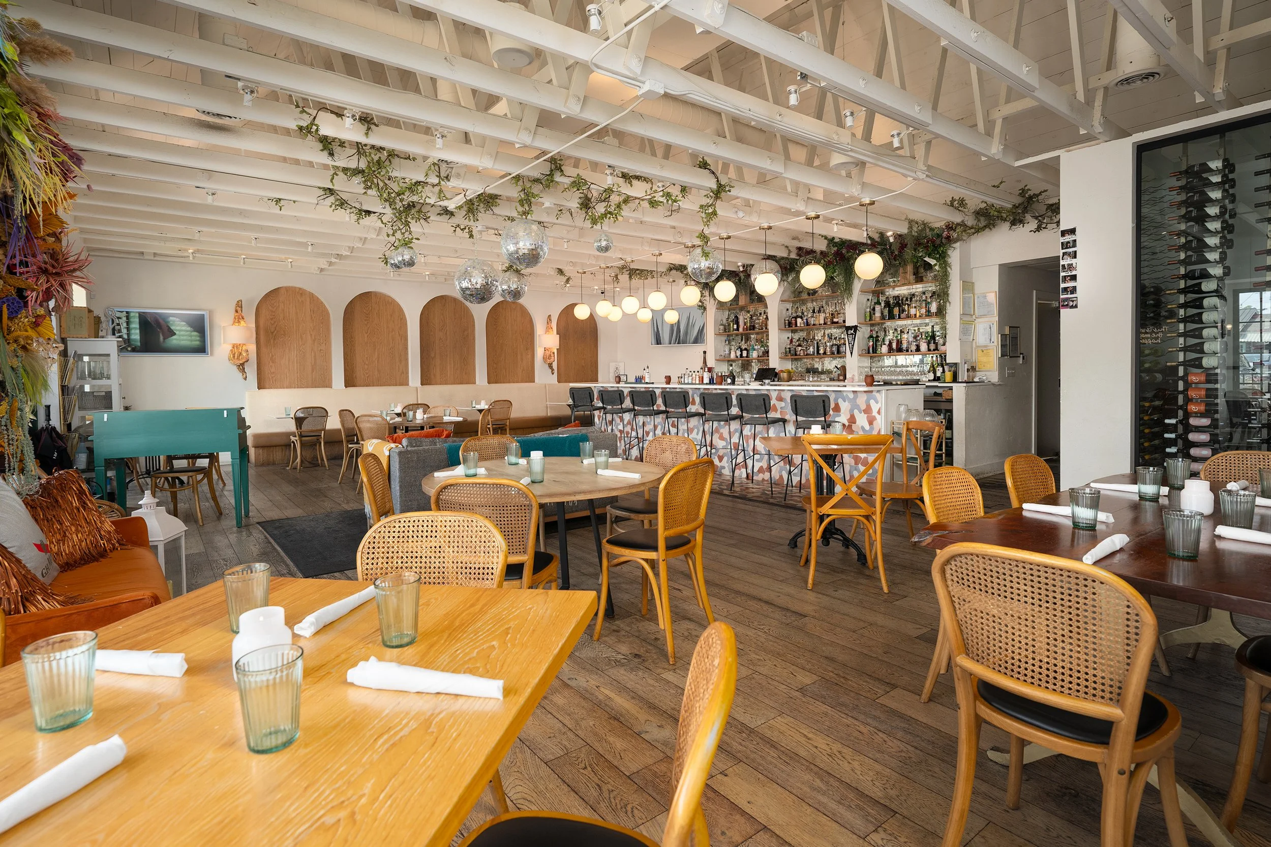 Interior of a restaurant with wooden flooring, tables, and chairs. The ceiling features exposed beams and hanging spherical lights. There is a bar with back shelves of alcohol bottles and bar stools. Decor includes green plants and disco balls hangin