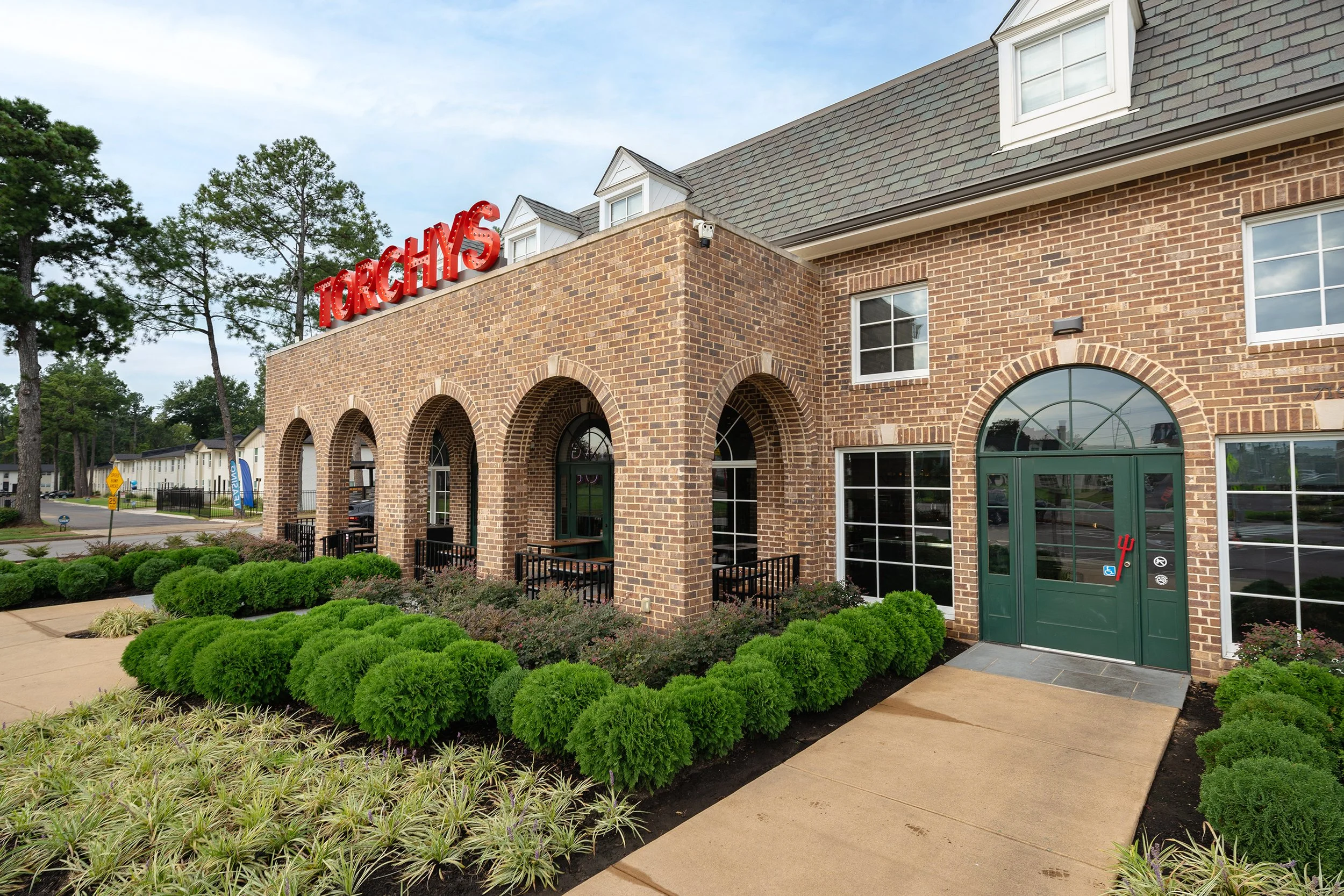 Exterior of a brick building with a neon sign that reads 'Torchy's' on the roof, green door with psi symbol stickers, arched windows, landscaped bushes and sidewalk in front.