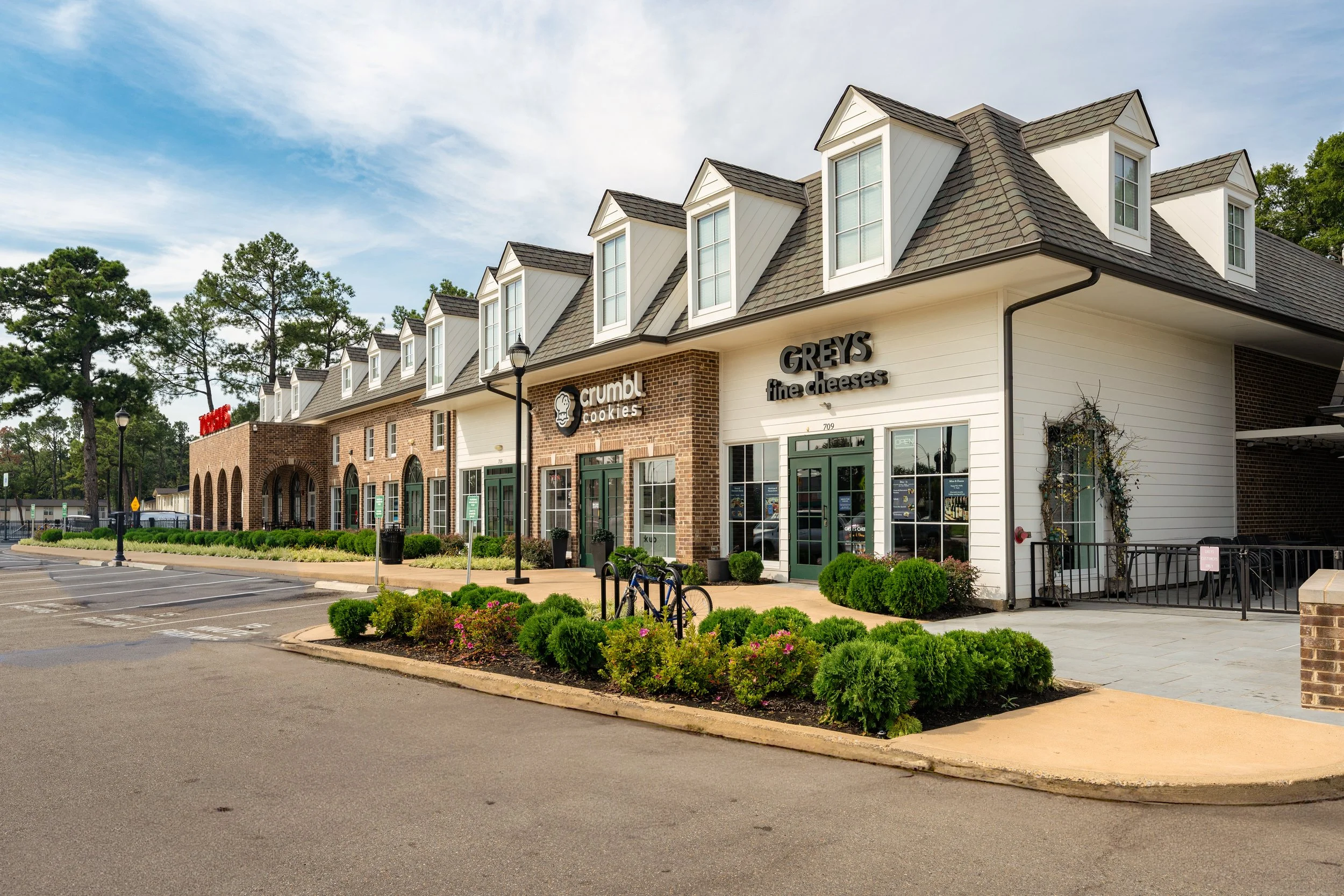 Shopping center with storefronts for Crumbl Cookies and Greys Fine Cheeses, landscaped sidewalk, parking lot, and cloudy sky.