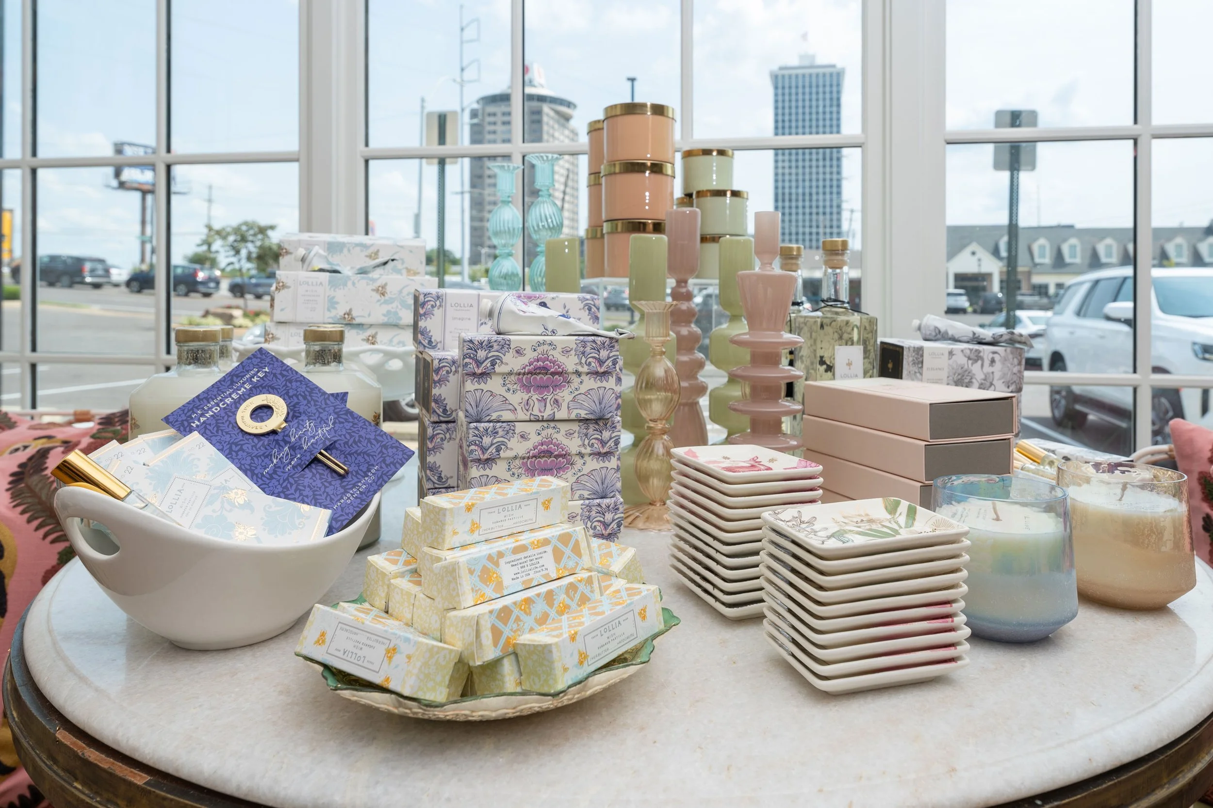 Display of various decorative candles, soap bars, and gift boxes on a table in front of a large window with a cityscape view.
