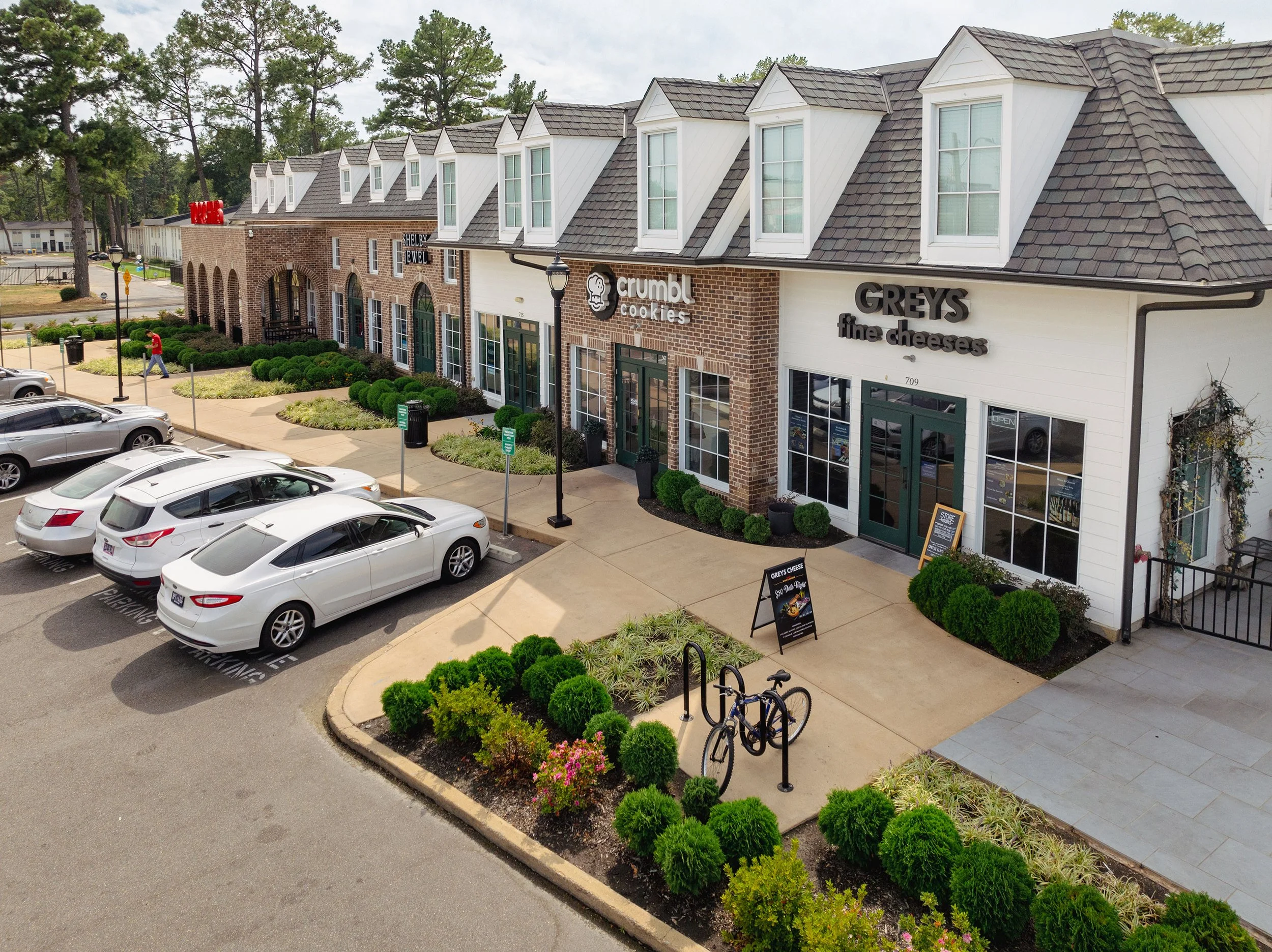A shopping complex with three visible storefronts, including Crumbl Cookies and Greys Fine Cheeses. There are parked cars in the parking lot and a woman walking on the sidewalk. The scene features landscaped bushes, a bicycle rack, and outdoor lighting.