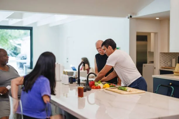 People gather around a kitchen island, preparing vegetables while an older man and a young man cook together, in a modern, bright kitchen.