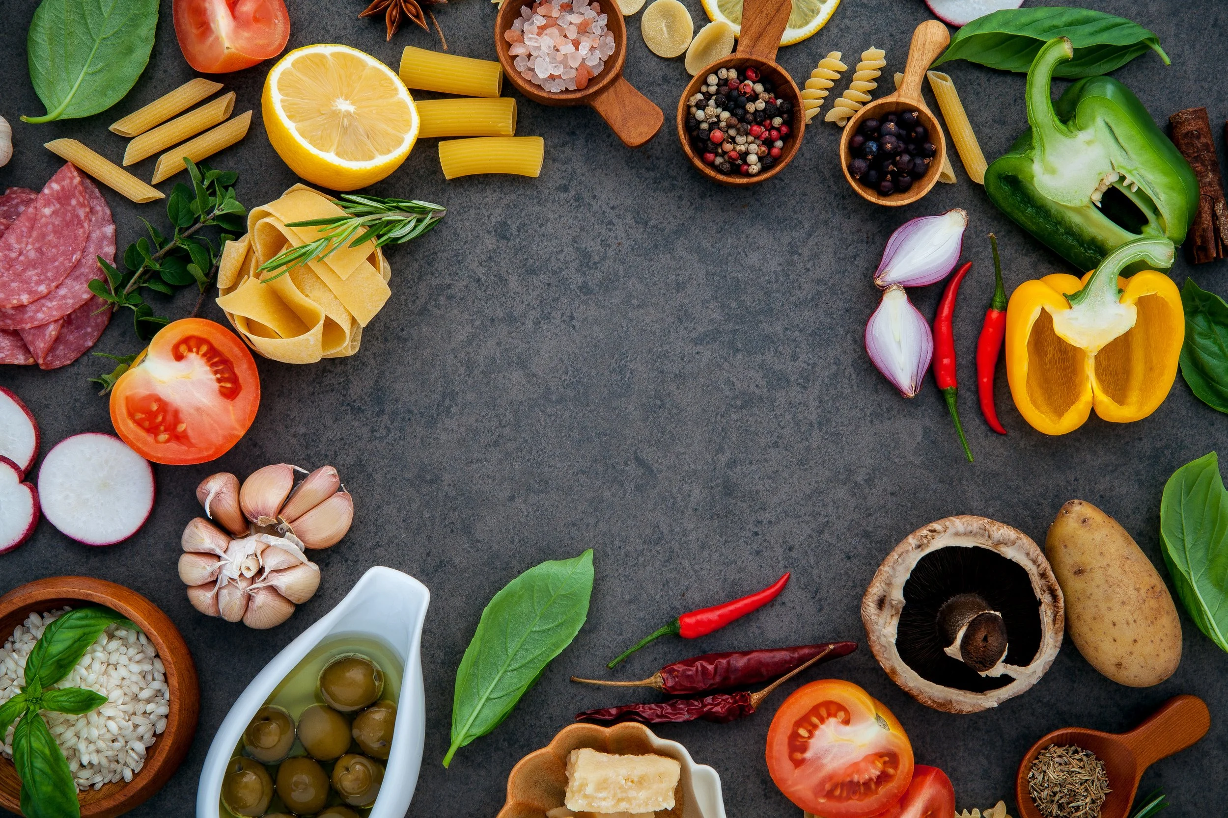 Assorted fresh vegetables, pasta, herbs, garlic, spices, and cooking ingredients arranged in a circle on a dark surface.