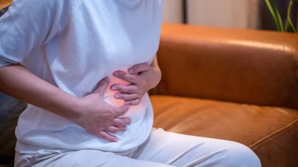 Person sitting on a brown leather couch, clutching their stomach with one hand, indicating abdominal pain.