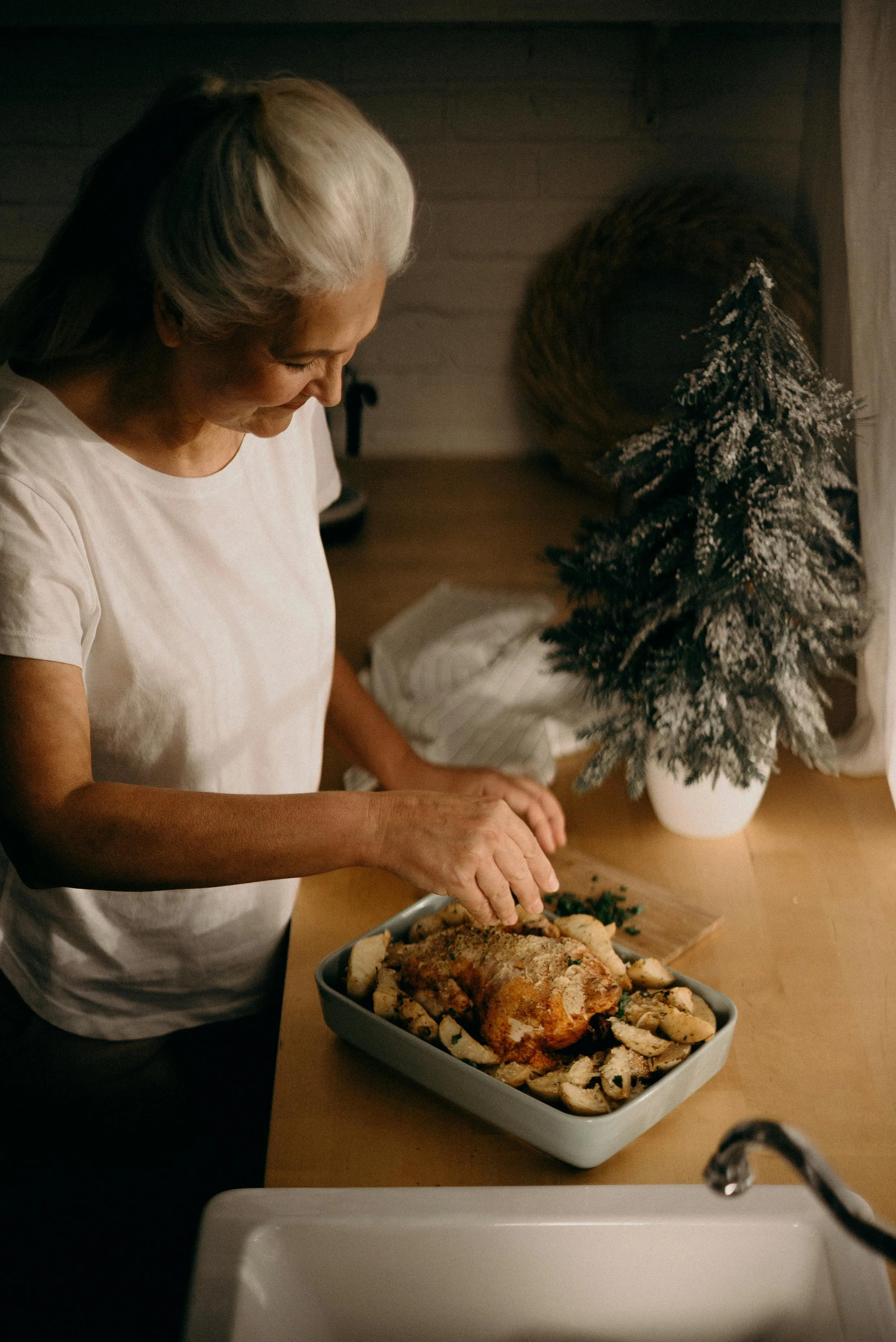 An elderly woman with gray hair standing at a kitchen counter, carving a roasted turkey with sliced potatoes and vegetables, decorated with a small snow-dusted Christmas tree in a white pot, in a cozy, warmly lit kitchen.