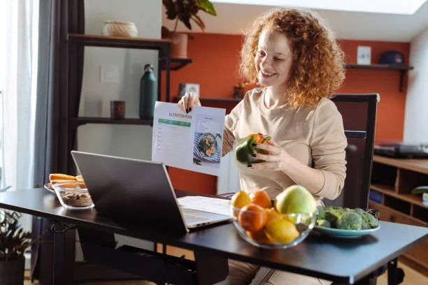 A woman with curly red hair smiling while sitting at a dining table with fruits and a laptop, reading a booklet.
