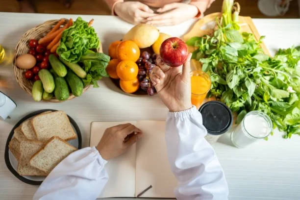 Overhead view of a person holding an apple above a white table with various fresh fruits, vegetables, bread, and drinks, with another person’s hands visible.