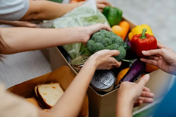 Multiple hands reaching into a box of fresh vegetables and canned goods, including peppers, broccoli, eggplant, and lettuce.