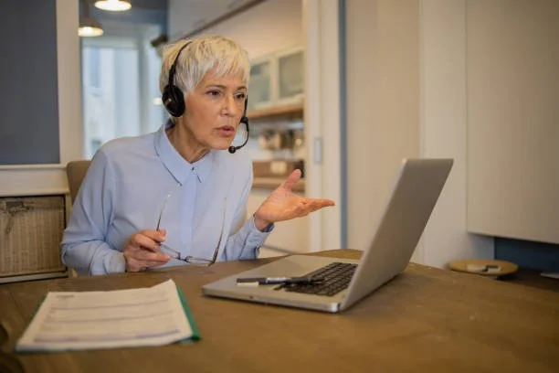 Older woman with short gray hair wearing a headset and light blue shirt, talking or questioning while looking at a laptop on a wooden table with papers and a pen.