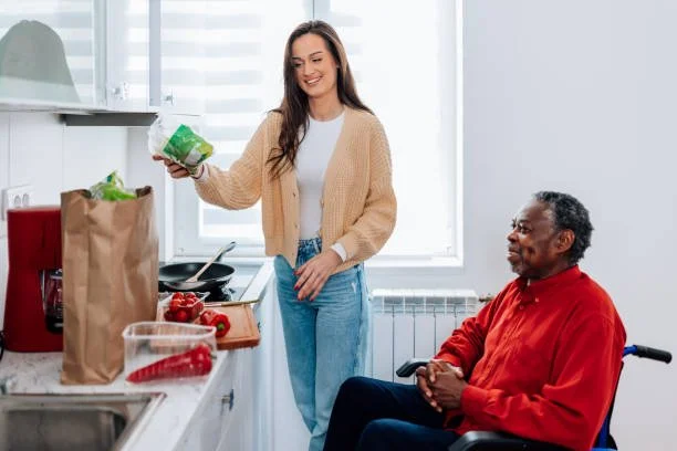 A woman and man in a kitchen with a window, with the woman holding a bag of grocery and the man sitting in a wheelchair, smiling.