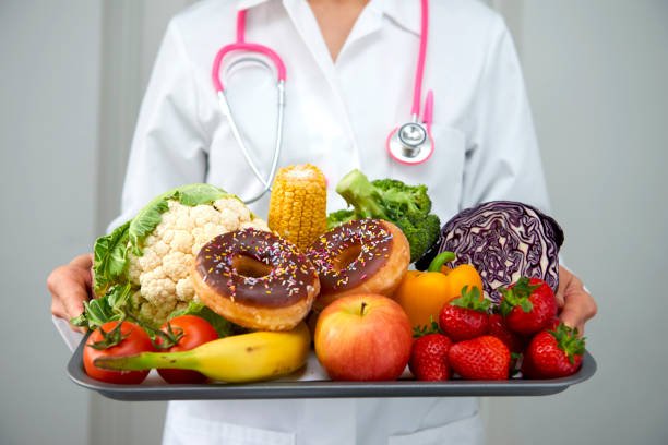 Person wearing a white coat and stethoscope holding a tray of fresh vegetables and donuts.