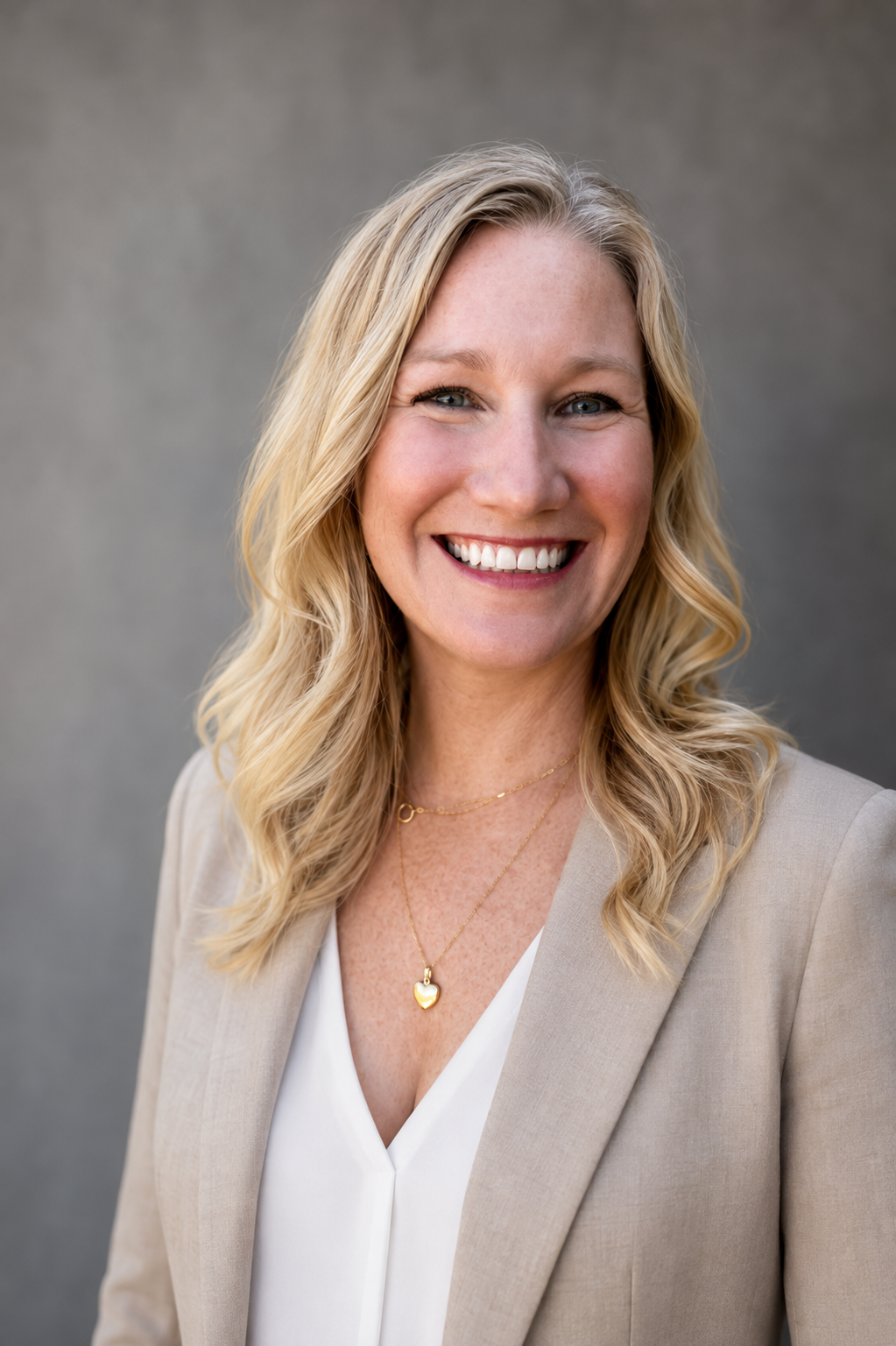 Portrait of a smiling woman with blonde hair, wearing a beige blazer and gold jewelry, standing against a neutral gray background.