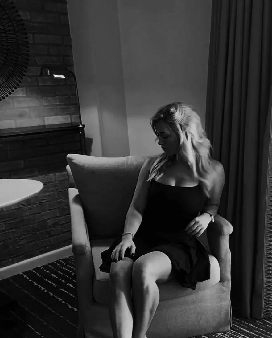 A woman with long wavy hair sitting on a cushioned armchair in a dimly lit room. She is wearing a black dress and looking down, with her hands resting on her lap and the arm of the chair.