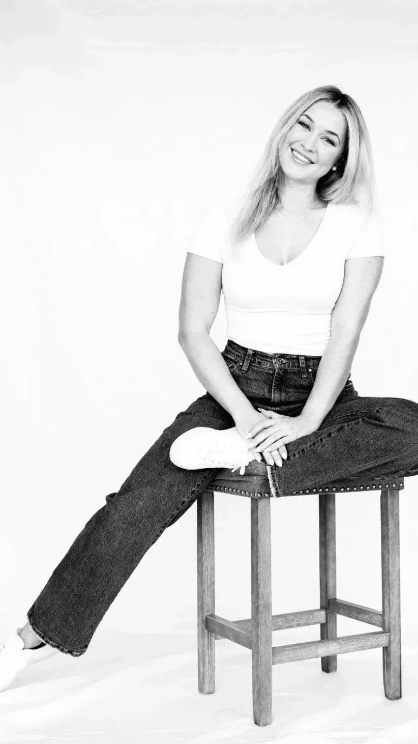Black and white photo of a woman sitting on a stool, smiling, wearing a white T-shirt and dark jeans, with her hair down.