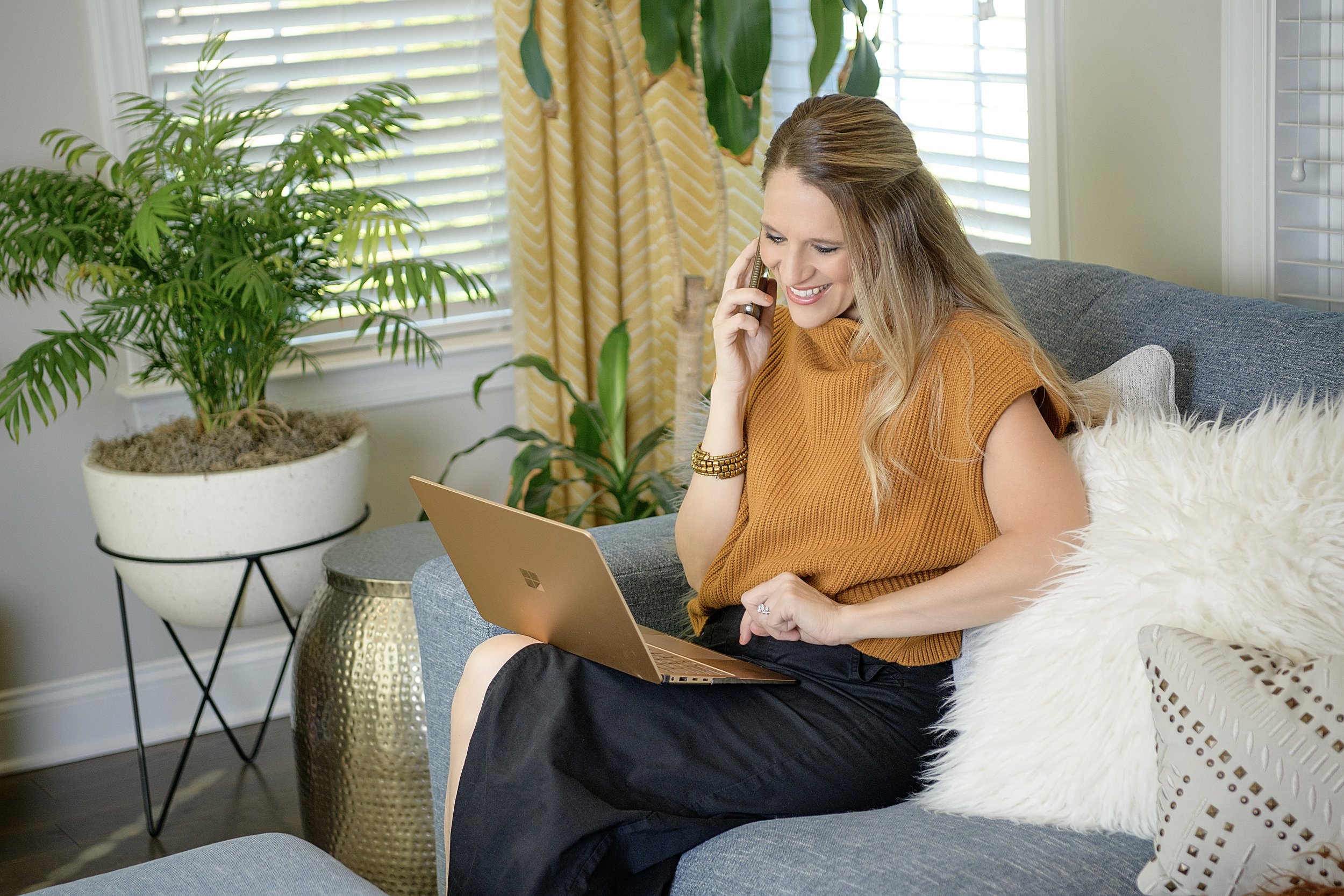 A woman sitting on a gray couch, talking on her cell phone, using a laptop, with plants and windows in the background.