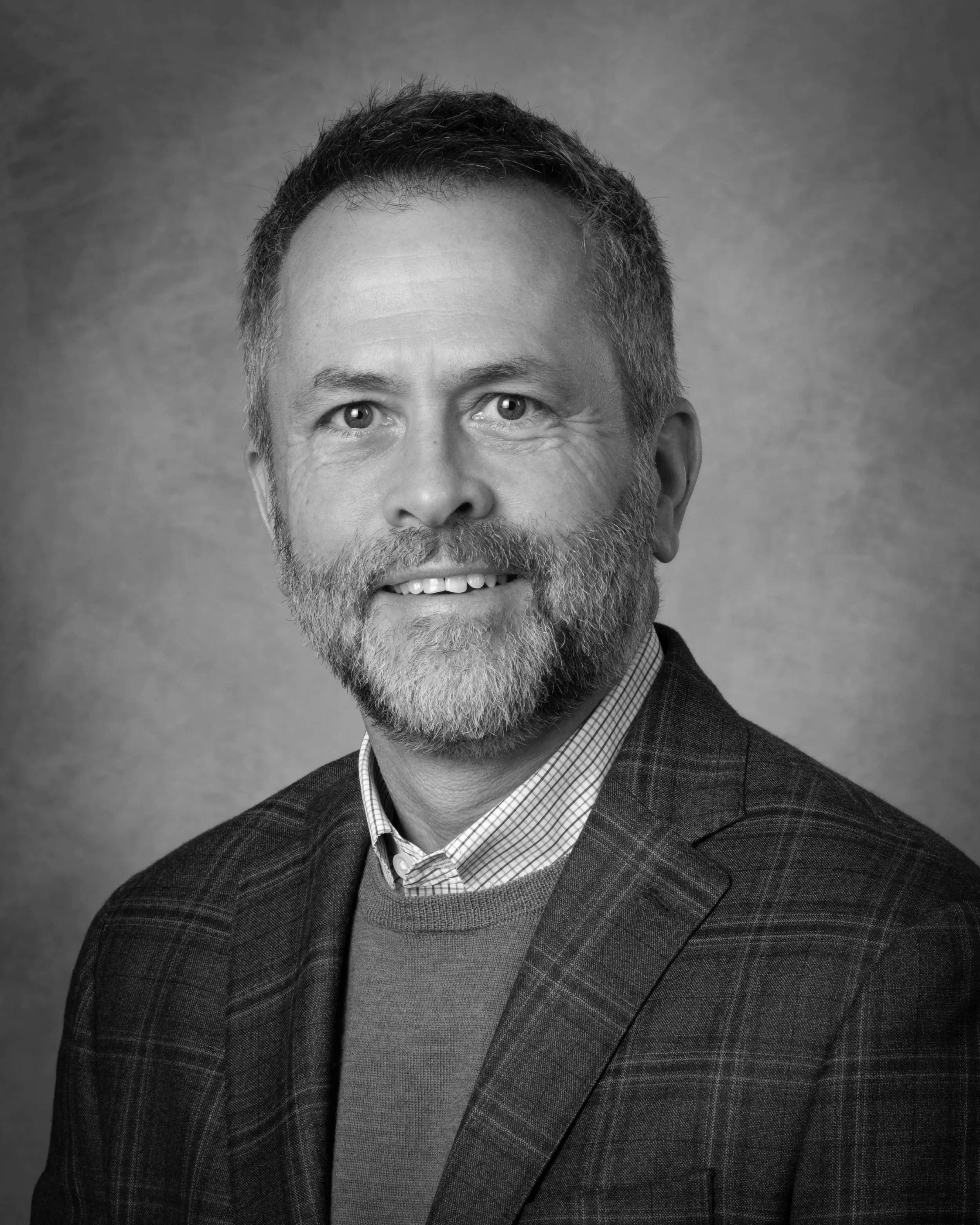 Black and white portrait of a man with a beard and short hair, wearing a checked dress shirt, a sweater, and a plaid blazer.