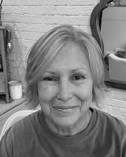 A smiling woman with short gray hair in a salon, with a mirror and hair salon supplies in the background.