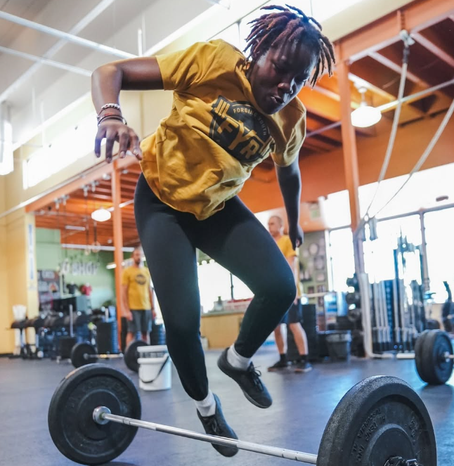 Young woman wearing a yellow t-shirt and black leggings balancing on a barbell in a gym during a workout.