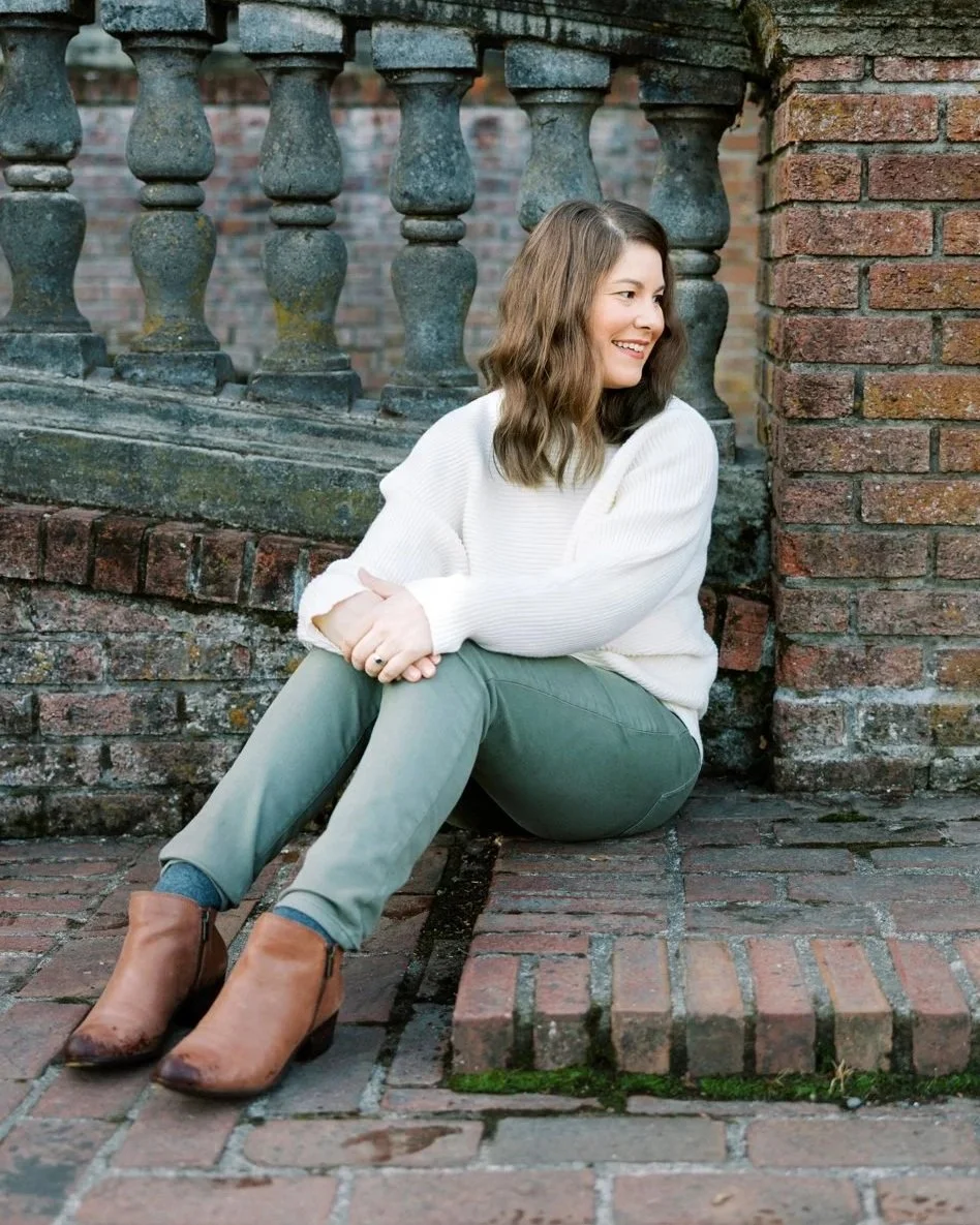 A woman with wavy brown hair sitting on a brick sidewalk, leaning against a brick wall with stone balusters in the background. She is smiling, wearing a white sweater, green pants, and brown ankle boots.