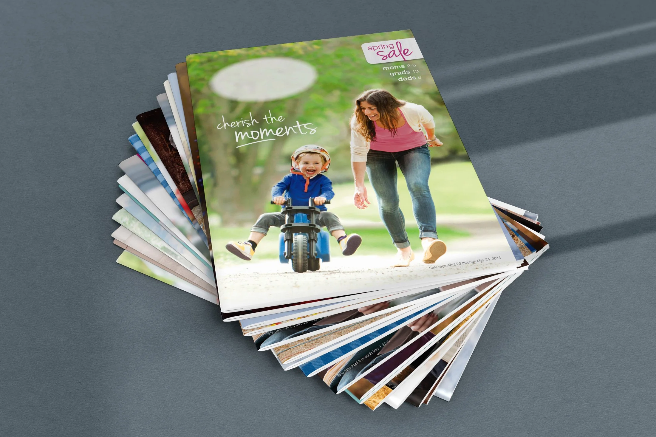 A stack of colorful magazines with the top magazine featuring a woman and a young boy riding a bike in a park, with the text 'cherish the moments' on the cover.
