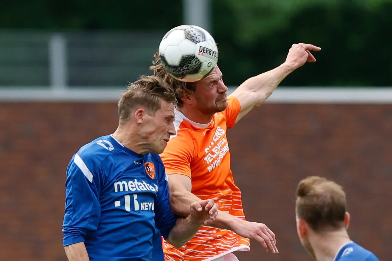 Twee voetbalspelers in actie tijdens een wedstrijd, één met een oranje jersey die de bal met het hoofd raakt en één met een blauwe jersey die probeert te verdedigen.